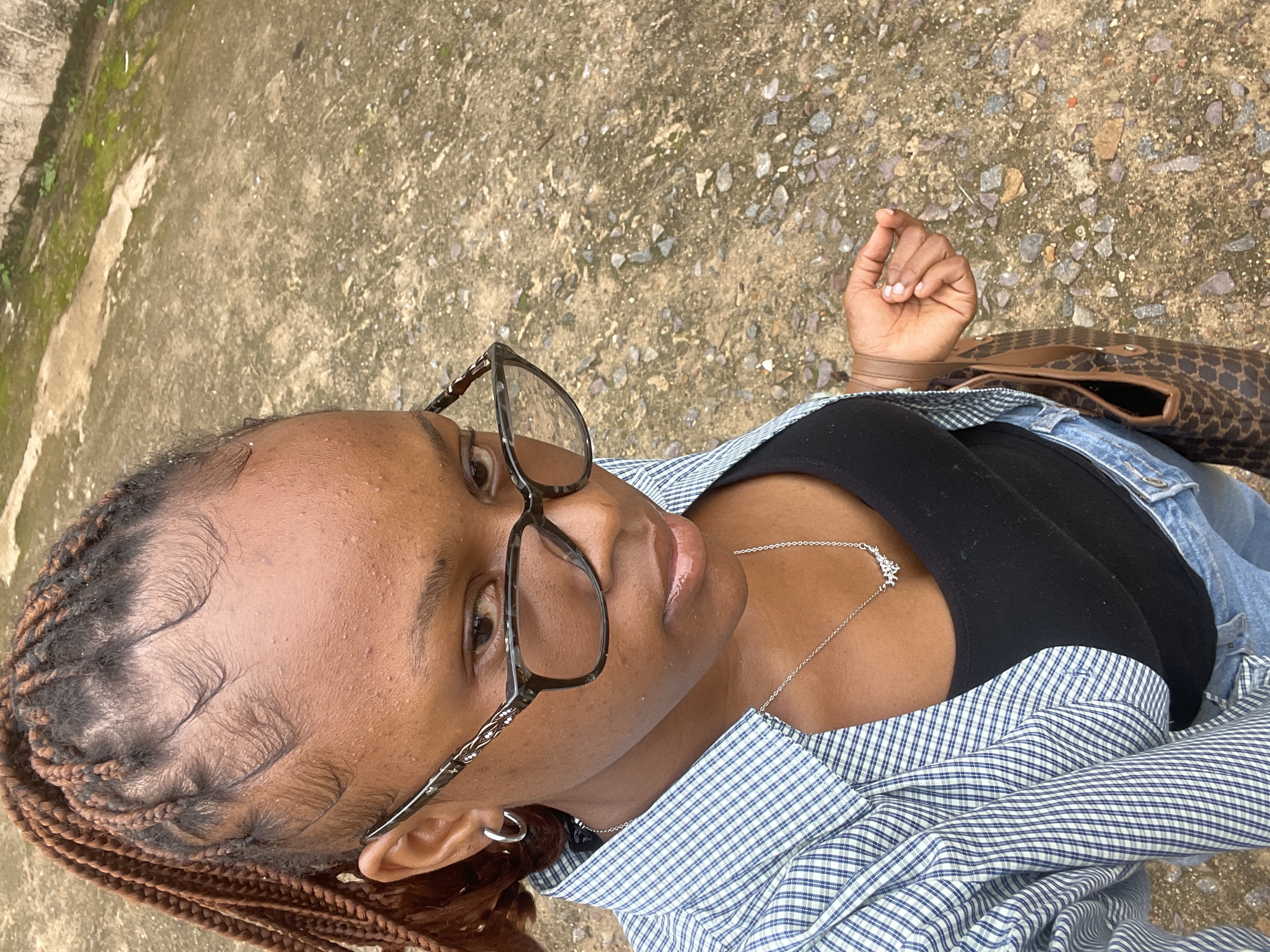 The media file shows a medium shot of a Black woman from a high-angle perspective, looking upwards and slightly to the right. She has braided hair, brown eyes, and is wearing glasses with a tortoiseshell pattern frame. She is also wearing a black tank top, a blue and white checkered shirt, and a silver necklace with a star-shaped pendant. A brown and black patterned handbag is visible on her right side. Her left hand is raised, with her index finger and thumb touching, and her right hand is resting on her lap, partially obscured by her clothing.
The background consists of a rough, concrete surface with scattered small stones and patches of green moss, suggesting an outdoor setting. The lighting indicates it might be daytime, possibly overcast due to the soft light. The overall mood appears relaxed or contemplative.
No visible text is present in the media file. The location context is Jos, Nigeria.