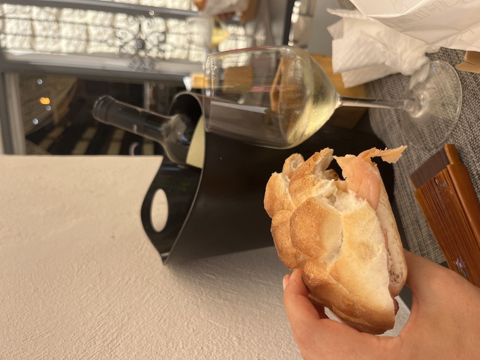 In Bari, Italy, a hand holds a sandwich in the foreground, with a glass of white wine and a bottle of wine in an ice bucket in the background. The scene is set against a textured wall and a table with a gray tablecloth, possibly at an outdoor dining area given the ambient lighting suggesting late afternoon or evening. A window with decorative grates is visible behind the wine bottle, hinting at the architectural style of Bari. The overall impression is a casual meal, perhaps an aperitivo, enjoyed in a restaurant or bar.