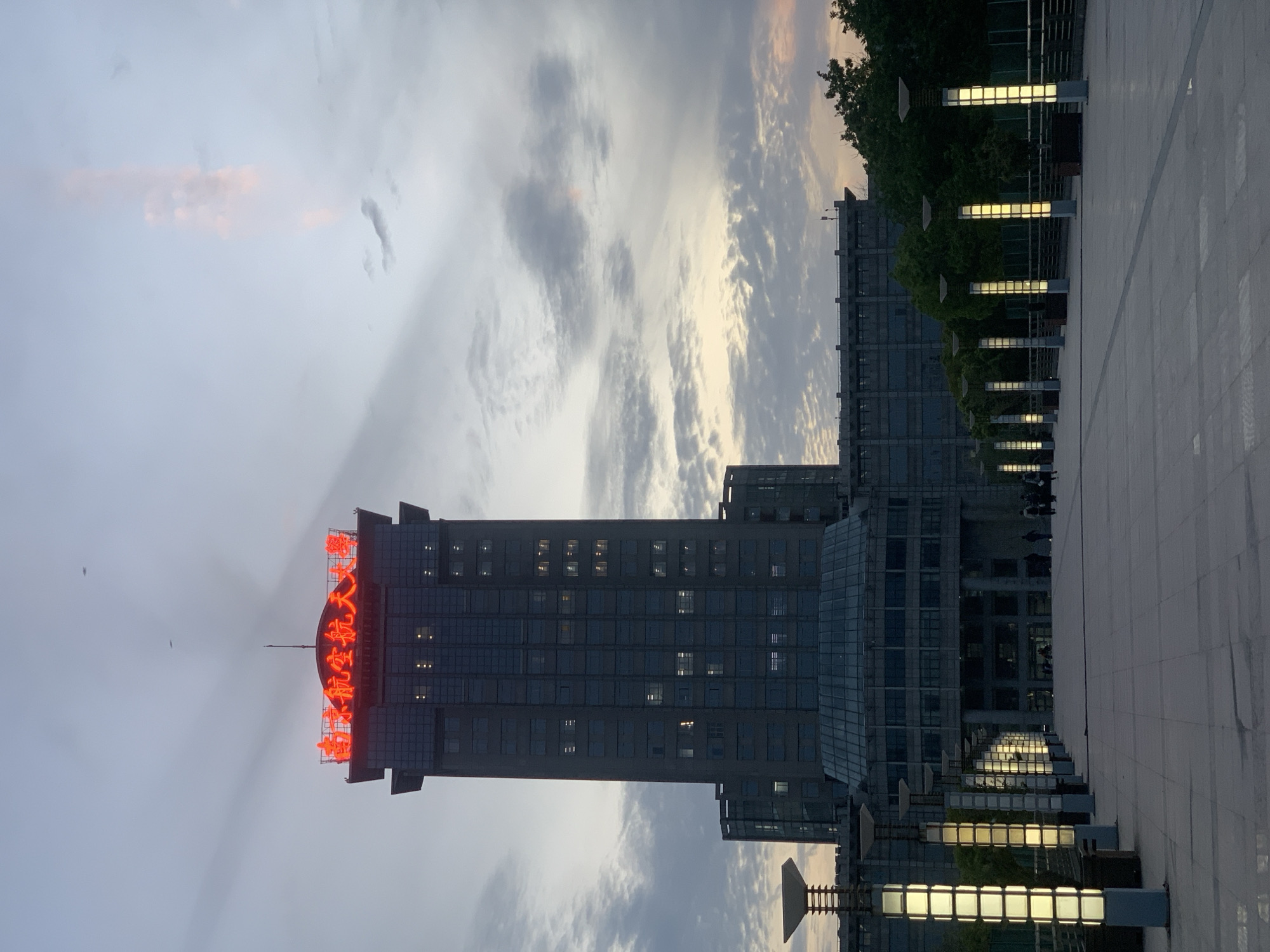 The image shows a tall building with Chinese characters in neon red lights on top. The building is located in Jiangning District, China, as indicated by the characters. The building's architecture is modern, with multiple floors and visible windows. The environment is urban, with other buildings and streetlights visible in the background. The sky is cloudy with some patches of sunlight. There are no people visible in the image. The building and the urban landscape are the central subjects.