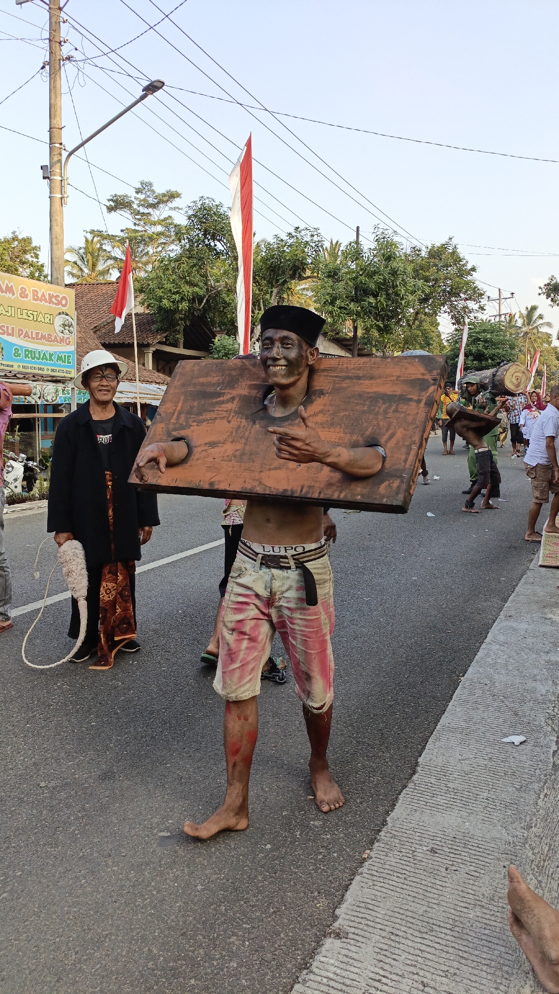 In Krajan Lor, Indonesia, a man wearing a black cap and shorts smeared with pink paint is participating in a parade. He is adorned with a large, wooden yoke that encircles his neck and shoulders, with holes for his arms. His face and body are covered in dark paint, and he is barefoot. He smiles and gestures with his right hand towards the camera. To his left stands an elderly person wearing a white hat, black jacket, and patterned skirt, holding a white rope with a fluffy end. In the background, Indonesian flags flutter, and trees line the street. Other parade participants are visible in the distance, some carrying logs. The event appears to be taking place on a paved road under a clear sky with utility poles and wires overhead.