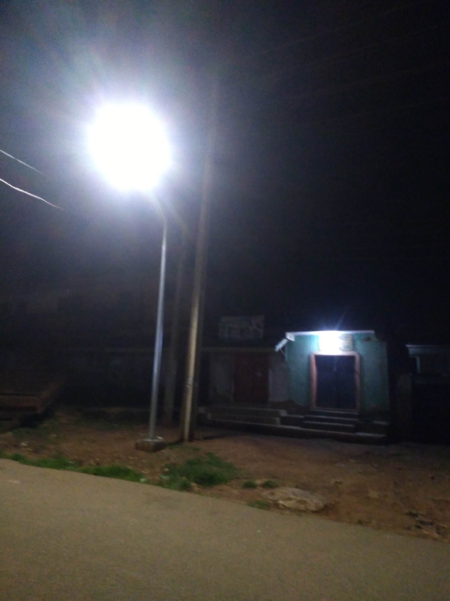 A night-time outdoor scene in Jos, Nigeria, captured with a bright street lamp illuminating the foreground and midground. The street lamp is a prominent feature, positioned slightly to the left of center, with its intense light creating a halo effect and washing out details in its immediate vicinity. Two poles for the street lamp are visible, with the one on the right appearing thicker and closer to the viewer.
In the midground, a small building with a green exterior and a doorway is visible. The doorway is dark, suggesting it is closed, and a wooden frame surrounds it. Above the doorway, a bright light emanates from within the building, illuminating the upper part of the structure and casting a glow on the steps leading up to it. To the left of this building, another structure with a red door is partially visible, also with steps leading to its entrance. The ground in front of the buildings is unpaved, appearing to be dirt and gravel with patches of dry grass. A paved road or walkway is in the foreground, curving to the left. The background is dark and indistinct, with a hint of structures or walls. The overall atmosphere is dimly lit and somewhat rural or semi-urban, typical of many areas in Jos at night. There are no people or animals visible in the scene.