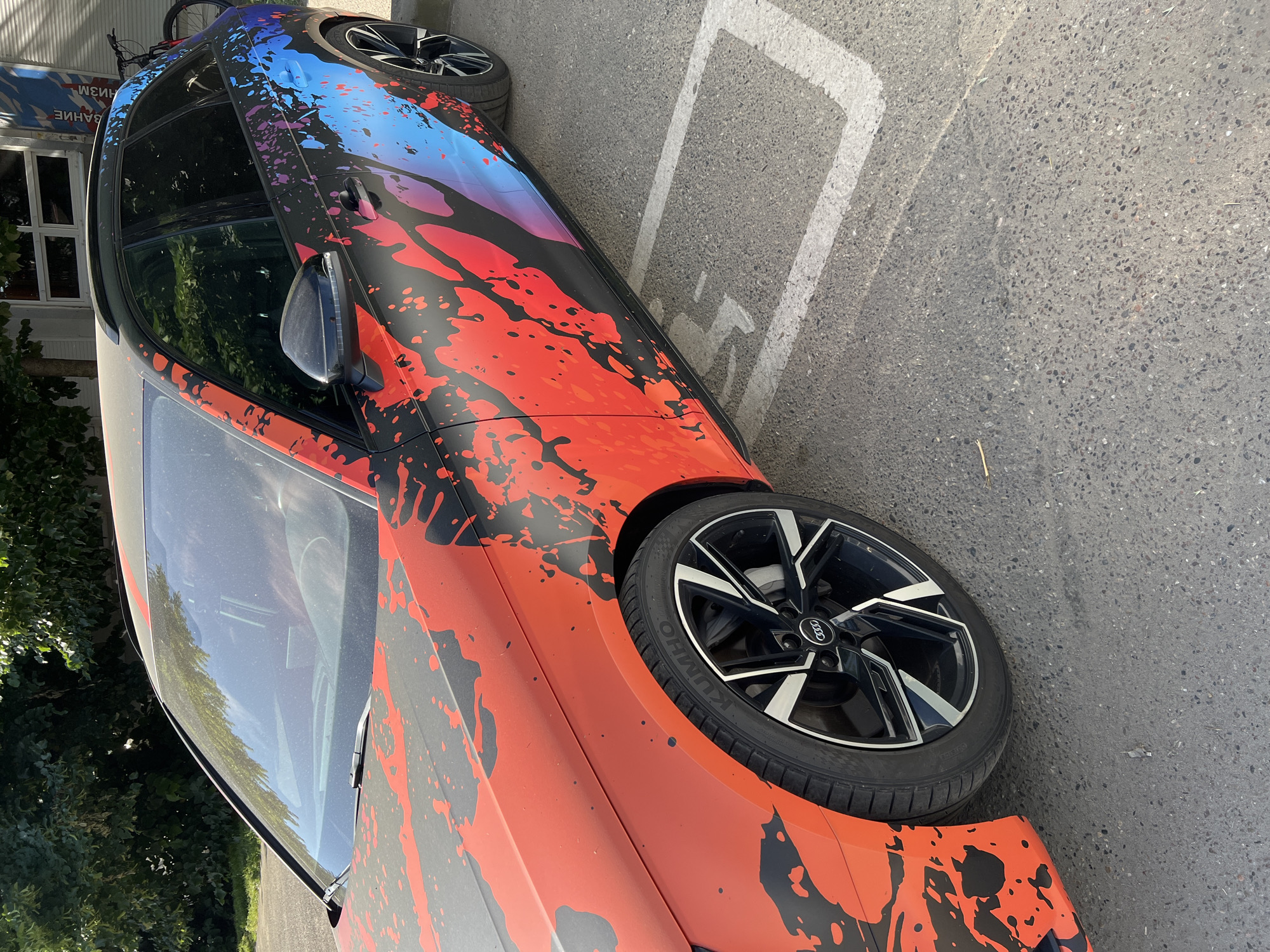 The image shows a close-up view of a black Audi station wagon with a vibrant red, black, and blue splatter graphic wrap parked on asphalt. The car is positioned in a parking spot marked with white lines, one of which is partially visible on the right side of the image. The front fender and wheel of the car are prominently featured on the left. The wheel is a black alloy with silver accents, and the tire is a Kumho brand. The car's paint is dusty. In the background, a tree with green foliage and a building with windows are visible. A bicycle is also visible in the background, leaning against the building. The scene appears to be an outdoor urban setting.