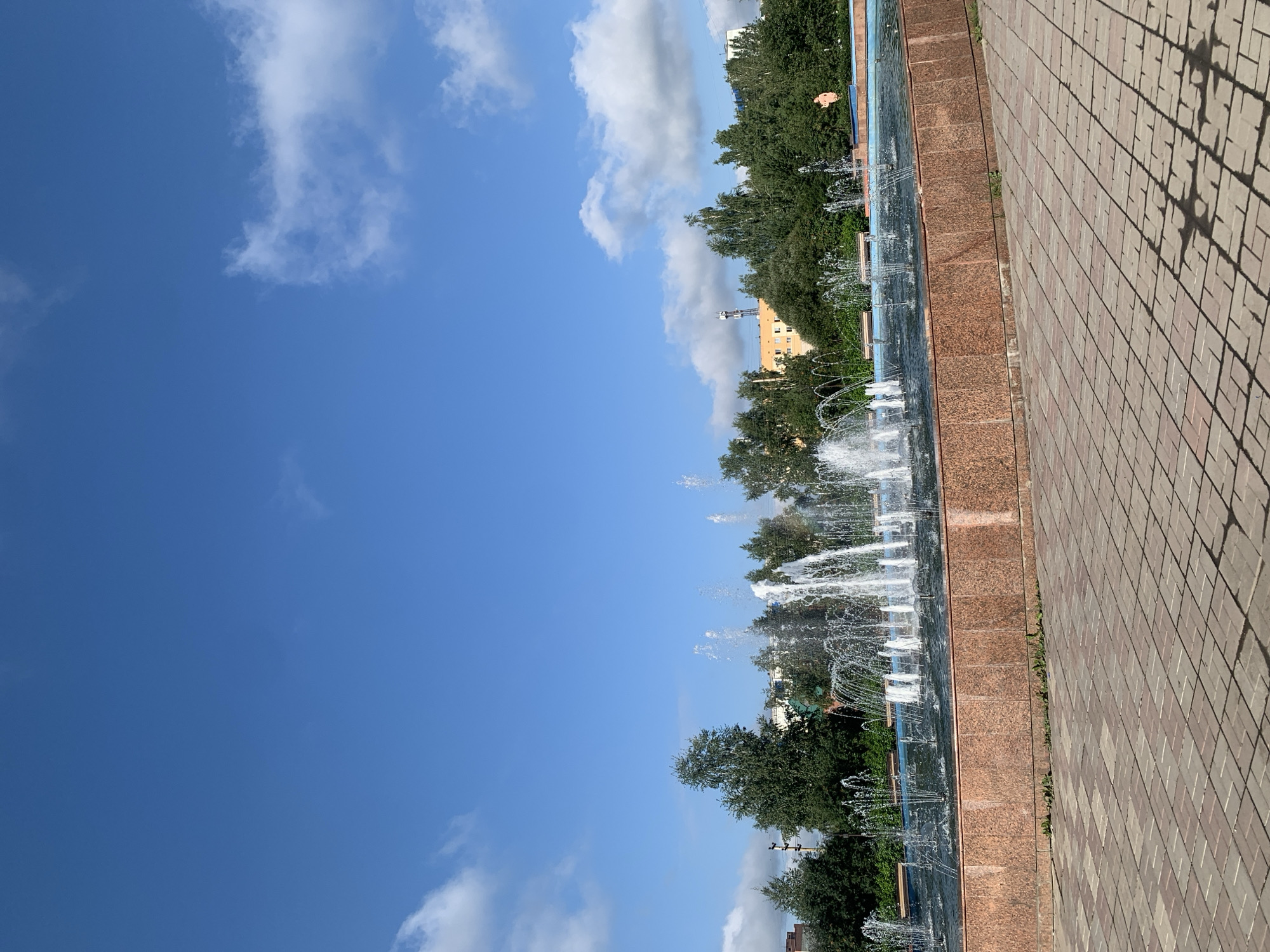 The image shows a large, multi-tiered fountain in an urban park setting. The fountain's water jets are active, creating a spray that catches the sunlight. The fountain is surrounded by a reddish-brown granite border. Beyond the fountain, there is a lush green line of trees, and further in the background, several buildings are visible, including what appears to be an orange-colored multi-story structure and a tower. The ground in the foreground is paved with brick-like tiles, casting shadows from an unseen light source, possibly trees or structures. The sky is a vibrant blue with scattered white clouds. A small, peach-colored statue of a person is visible on the far right edge of the fountain's basin. This scene is located at the fountain complex on Novyy Gorod Street in Prokopyevsk, Russia.