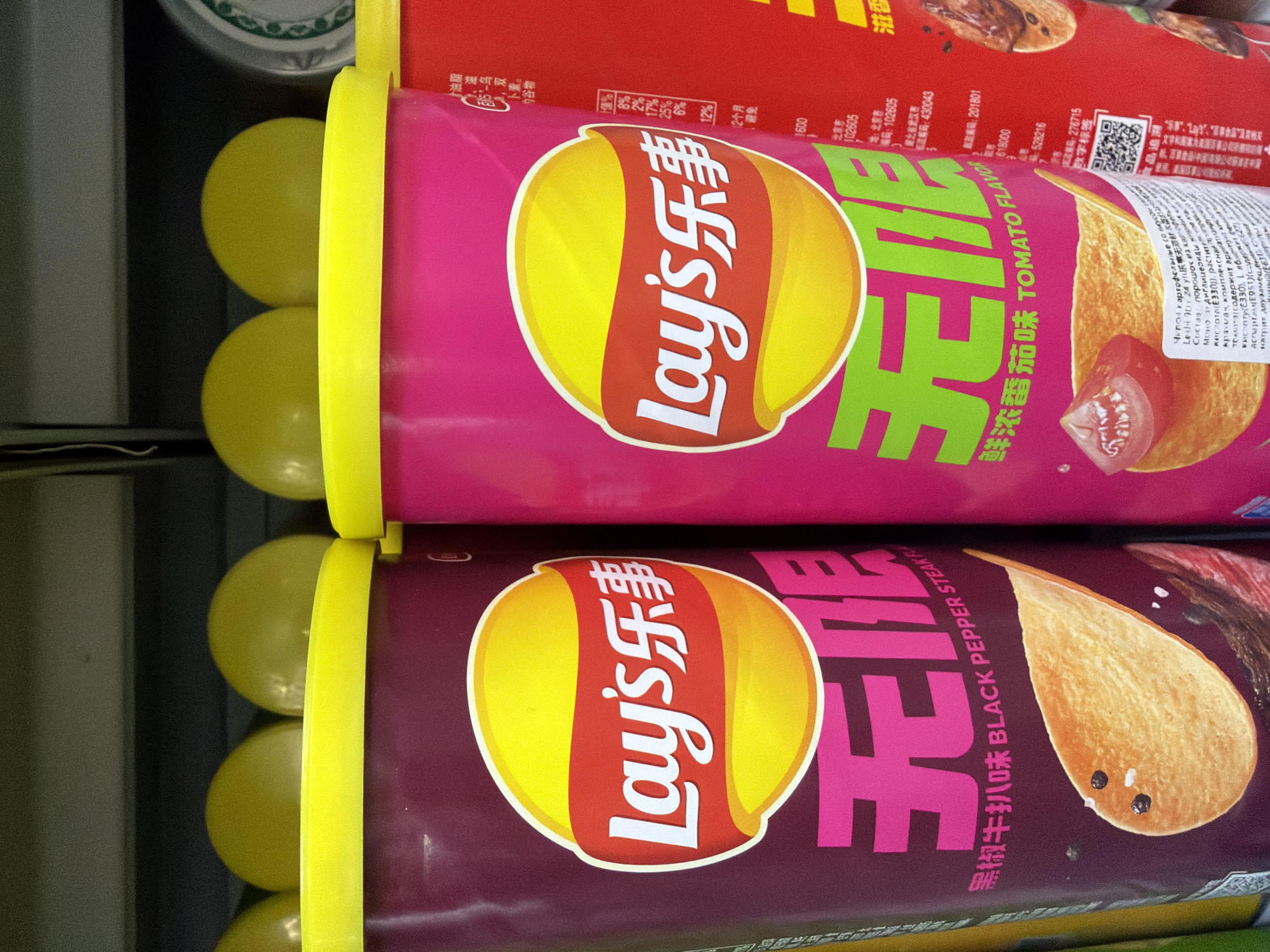 The image shows two cans of Lay's potato chips stacked vertically on a shelf. The top can is pink and has the flavor "Tomato Flavor," while the bottom can is dark purple and has the flavor "Black Pepper Steak Flavor." Each can has a yellow lid and the Lay's logo prominently displayed. The text on the cans is in both English and Chinese. The cans are surrounded by other items in a retail setting, most likely a supermarket. Some of the text on the cans references locations such as Beijing. Moscow, Russia is not mentioned in the text or depicted in the image.