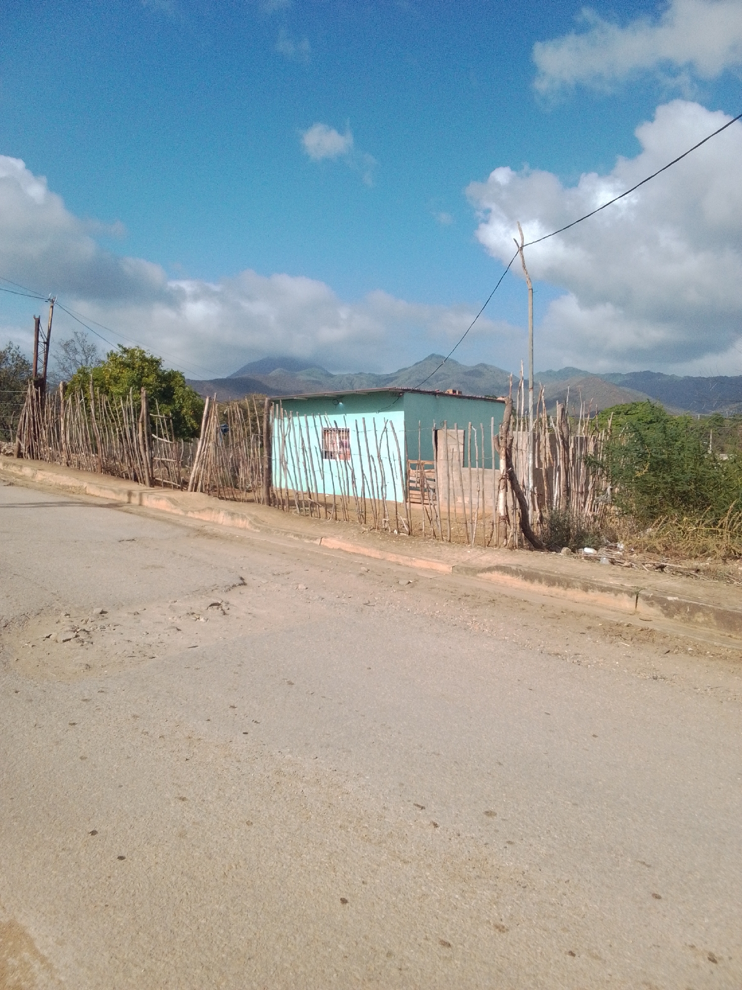 The image depicts a rural or semi-rural scene in La Tagua, Venezuela, under a bright blue sky with scattered white clouds. In the foreground, a dirt road curves upwards, showing signs of wear with patches of loose gravel and dark spots. Along the right side of the road, a simple, light blue house with a reddish-brown window is partially visible, surrounded by a fence made of vertically placed, rough wooden poles. This fence extends to the left, partially enclosing an area with a rustic wooden gate and some vegetation. Beyond the fence, a line of trees with green foliage is seen, and in the background, rolling hills and mountains are silhouetted against the sky. A utility pole with power lines is also present, adding to the sense of infrastructure in the landscape. The overall impression is one of a modest dwelling in a natural setting, with evidence of basic construction and the surrounding environment.