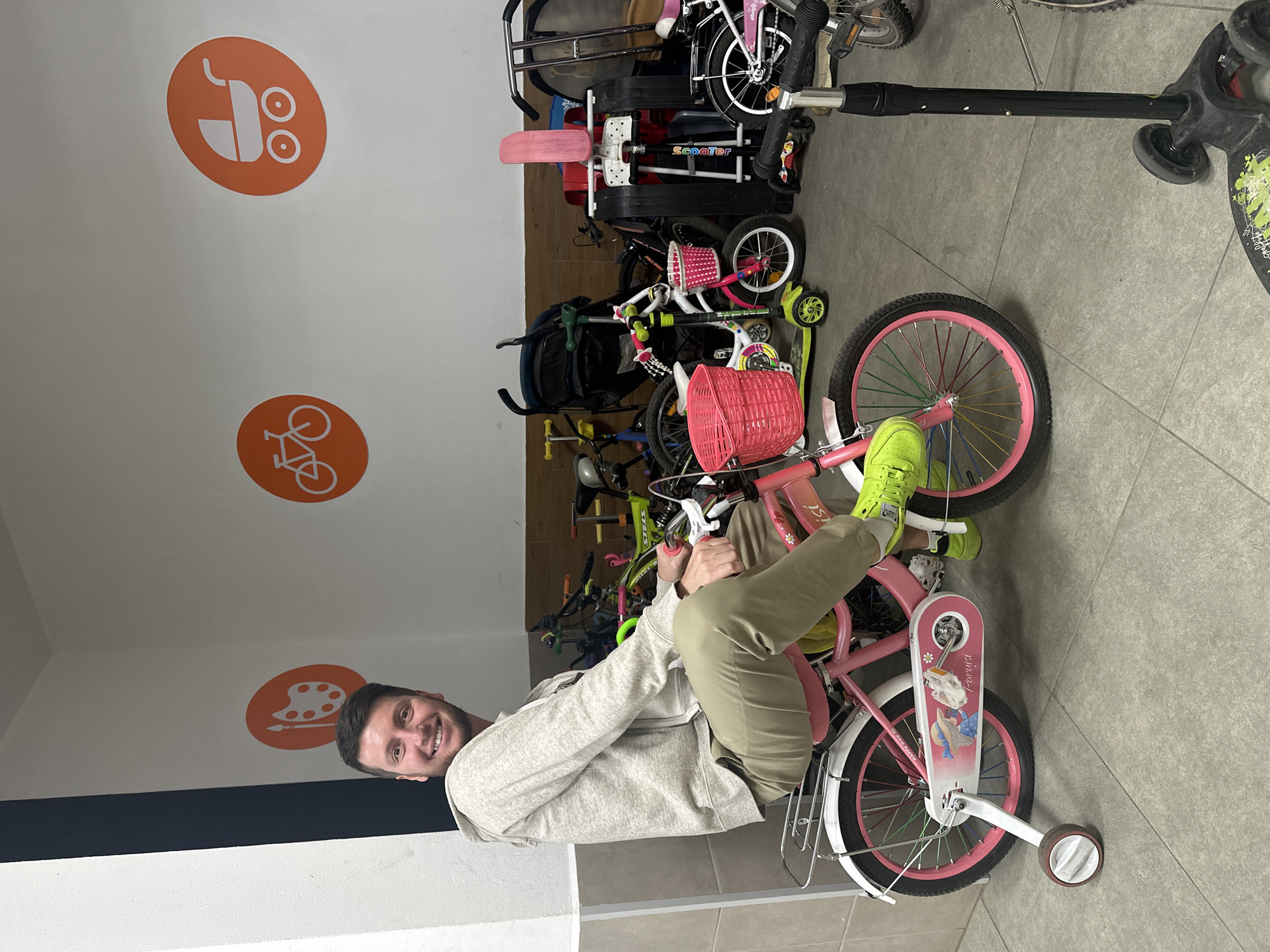 A man is sitting on a child's pink bicycle with training wheels, indoors. The man, wearing a grey hoodie and khaki pants, is smiling at the camera. The bicycle has a pink basket on the handlebars and a decorative sticker on the chain guard. The background shows a collection of other bicycles, scooters, and a stroller, suggesting a storage area or a shop. On the wall are orange circular stickers with white icons of a palette and paintbrush, a bicycle, and a baby carriage. The floor is tiled. The location is specified as Мирный, Russia.