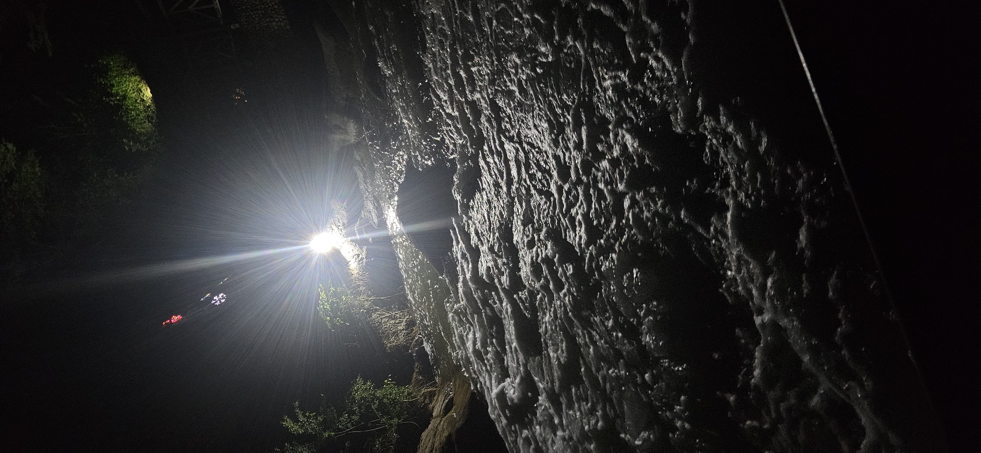 A brightly lit, dark nighttime scene is presented with a focus on a craggy, textured surface, likely a rock face or heavily textured wall, to the right. The texture is accentuated by the strong light source positioned to the left of the frame, casting stark shadows and highlights. The light creates intense glare and multiple light rays that shoot out in all directions. There are what appear to be lights in the background. Some green foliage is visible at the bottom and slightly above the light source. The setting appears to be outdoors at night, and due to the lack of other indicators, the specific location within the city of Unknown, Unknown, is not discernible.