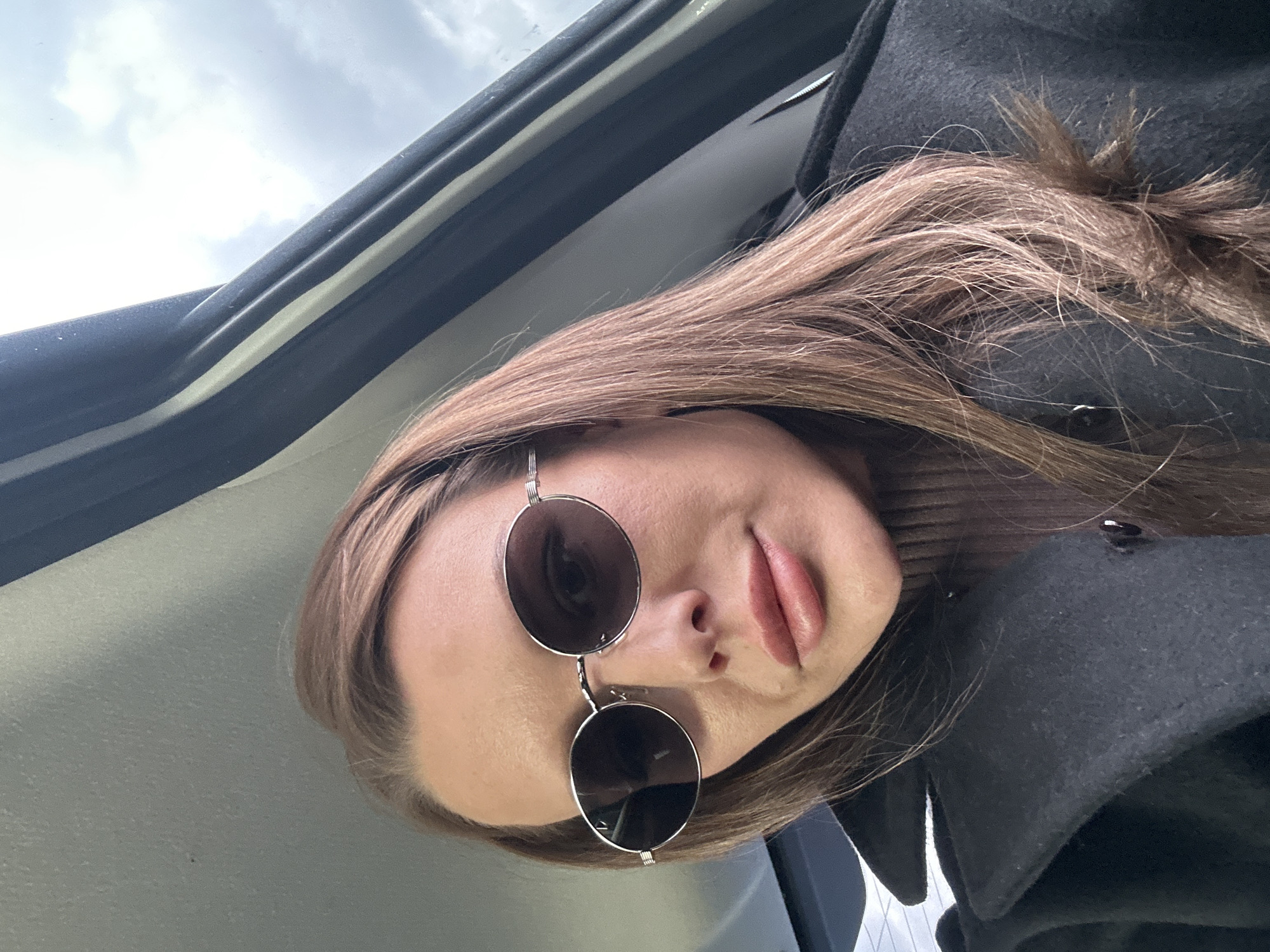 A woman is posing for a selfie from inside a vehicle. She is wearing round sunglasses with silver frames and a dark coat over a brown turtleneck sweater. Her long, brown hair is slightly disheveled. The interior of the vehicle is visible, showing the roof and part of the dashboard. The background shows a cloudy sky. The exact location in the city of Chaikovsky, Russia is not discernible from the image.