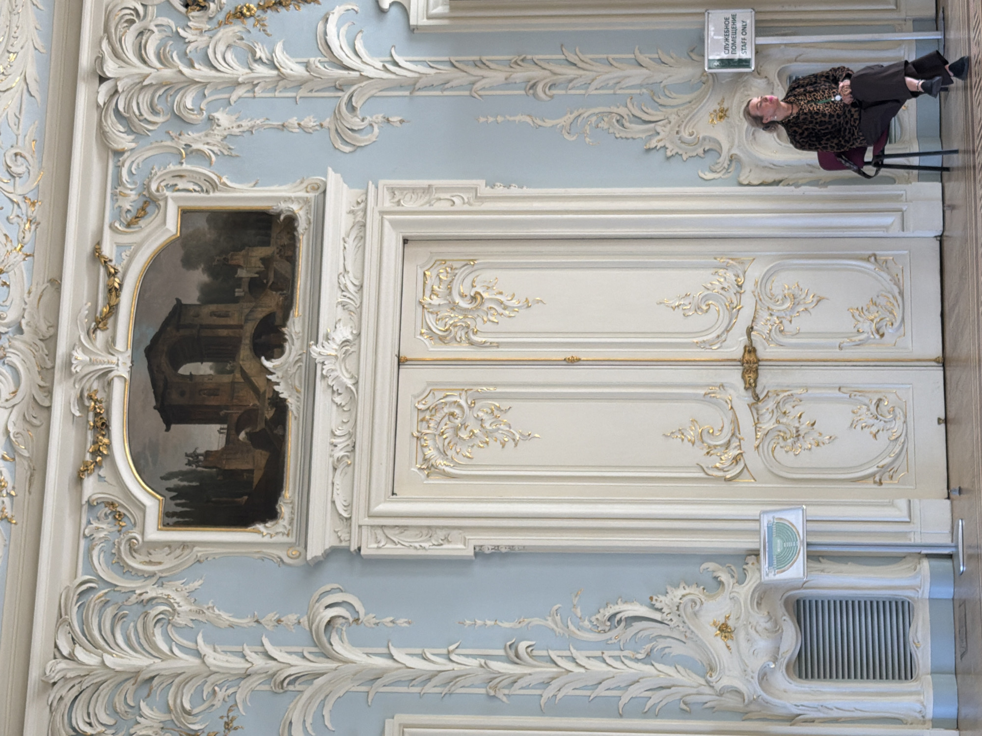 The image depicts the opulent interior of a grand building, likely a palace or museum, in Saint Petersburg, Russia. The walls are painted a soft blue and adorned with elaborate white and gold baroque-style stucco work, featuring intricate floral and scroll motifs. In the center of the scene are a pair of tall, double doors, also painted white with gold detailing. To the left of the doors, a decorative fireplace surround houses a modern-looking radiator grille. A small, silver-colored stand with a screen displaying a green diagram is placed in front of the fireplace. On the right side of the doors, a woman is seated on a dark red chair, facing away from the camera and towards the doors. She is wearing a black and brown spotted jacket over a patterned shirt and dark trousers. Next to her, a white sign with green lettering, stating "СЛУЖЕБНОЕ ПОМЕЩЕНИЕ STAFF ONLY," is mounted on a pole. The floor appears to be polished wood. Above the doors, a rectangular painting in an ornate gold frame depicts a classical architectural scene with arches and figures. The overall impression is one of historical grandeur and meticulous decoration.