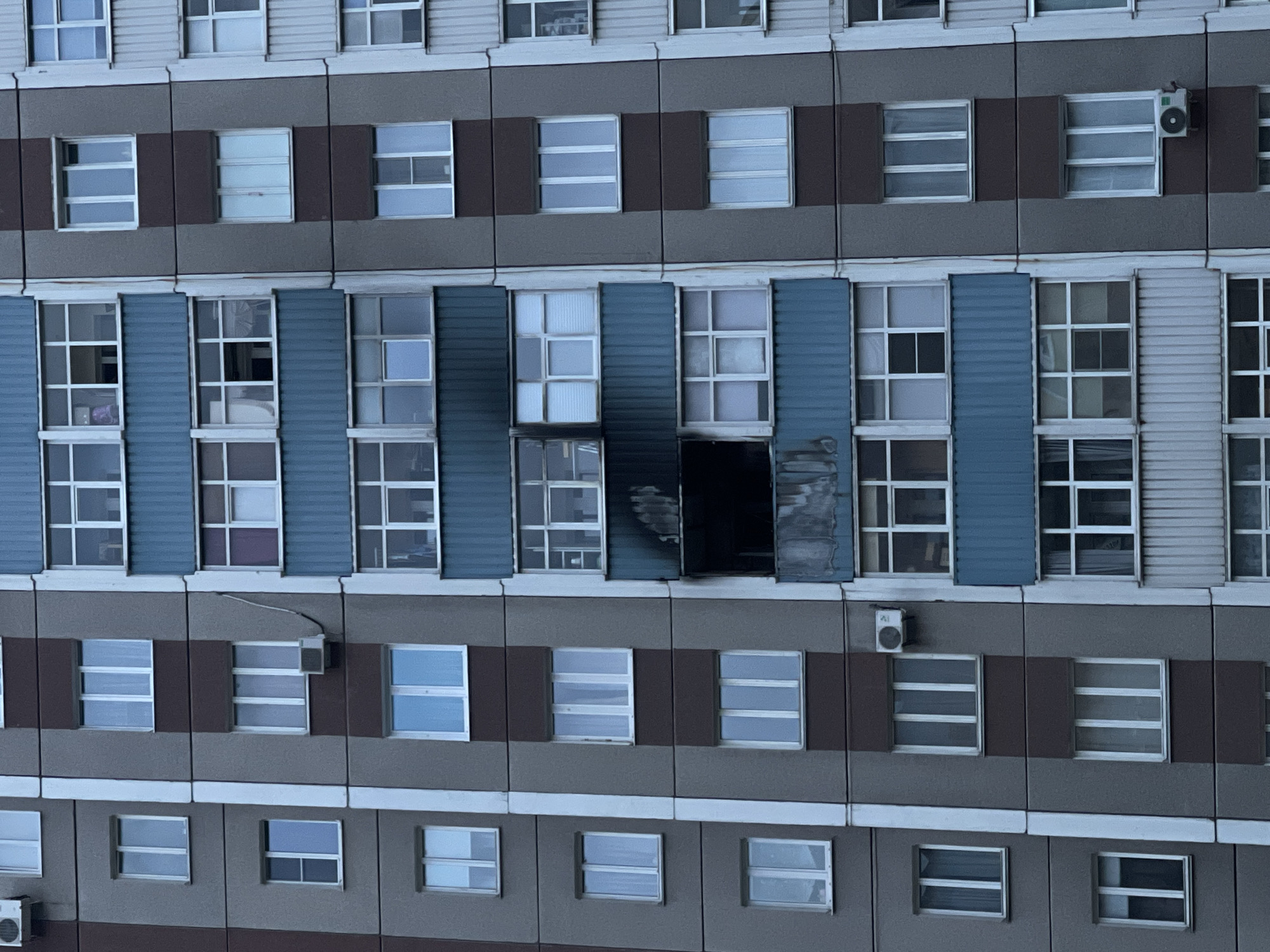 A tall, grey apartment building is shown in a vertical, full-frame shot. The building has multiple stories, each with several windows and balconies. Some balconies are enclosed with glass, while others are open. The building has a predominantly grey facade with some brown and blue accents. On one of the balconies, there are signs of fire damage, with blackened areas and debris visible. The windows are mostly glass panes in white frames. The image appears to be taken during the daytime, with overcast skies. There are no people visible in the image.