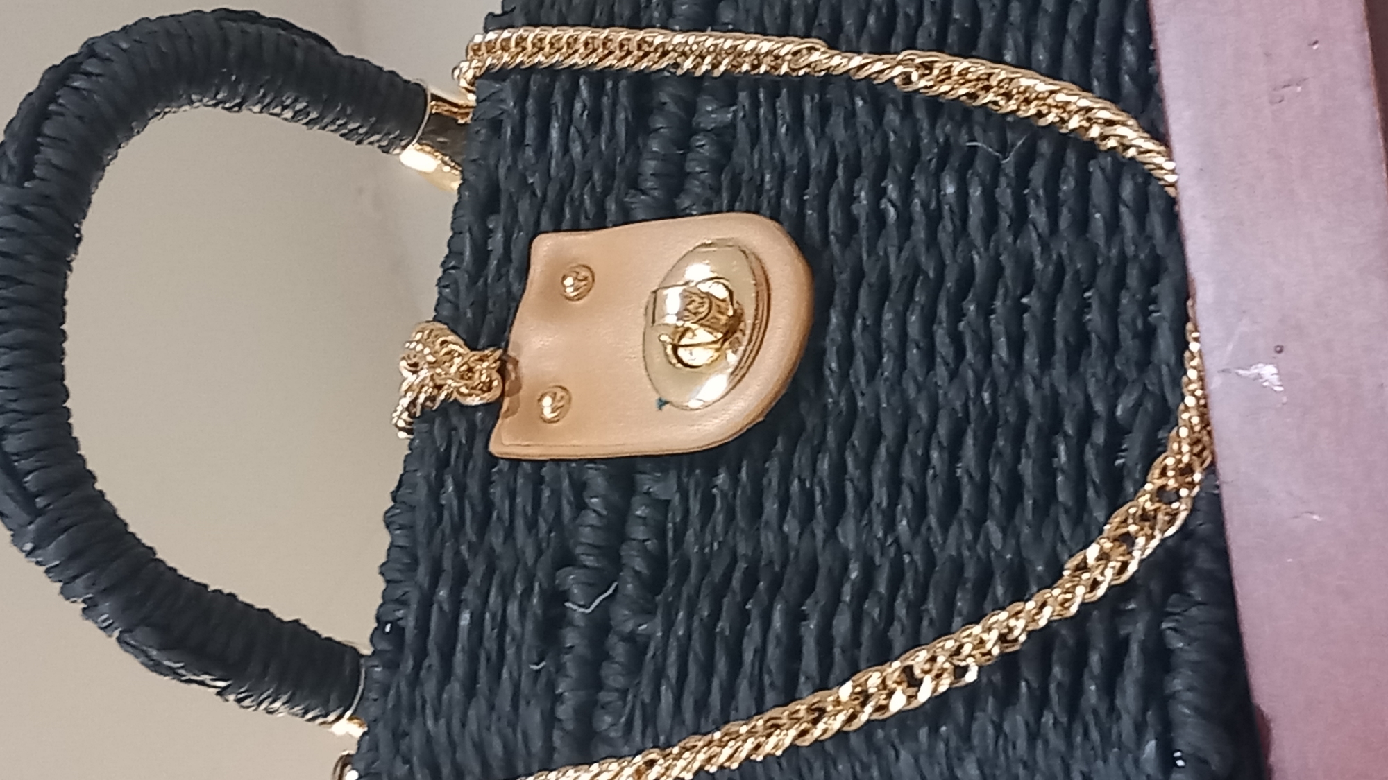A close-up, eye-level shot shows the top portion of a black woven handbag. The bag has a thick, rounded handle made of the same black woven material. A gold-colored metal chain is draped across the front of the bag, curving down from the top left and then across the bottom right of the frame. On the front of the bag, just below the handle, is a decorative element: a tan, shield-shaped piece of material with two small gold studs resembling eyes and a gold clasp at the bottom. This embellishment suggests a whimsical design, possibly for a women's accessory. The background is a plain, light-colored surface, out of focus. The image is taken indoors, and there is no visible indication of the city of Kos, Greece.