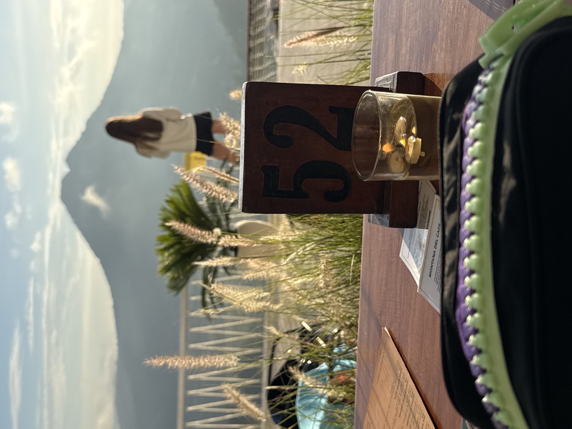 The image, taken in Kedisan, Indonesia, shows a partially obscured table at a restaurant with a view of a mountain. The table has a wooden stand with the number "52" displayed, a glass with decorative elements inside, a menu, and a black bag with purple and light green details. In the background, beyond a railing with decorative grasses, a woman wearing a white top and dark shorts is visible. A palm tree is to the left of her and in the far background is the large mountain. The sky is light and slightly overcast.
