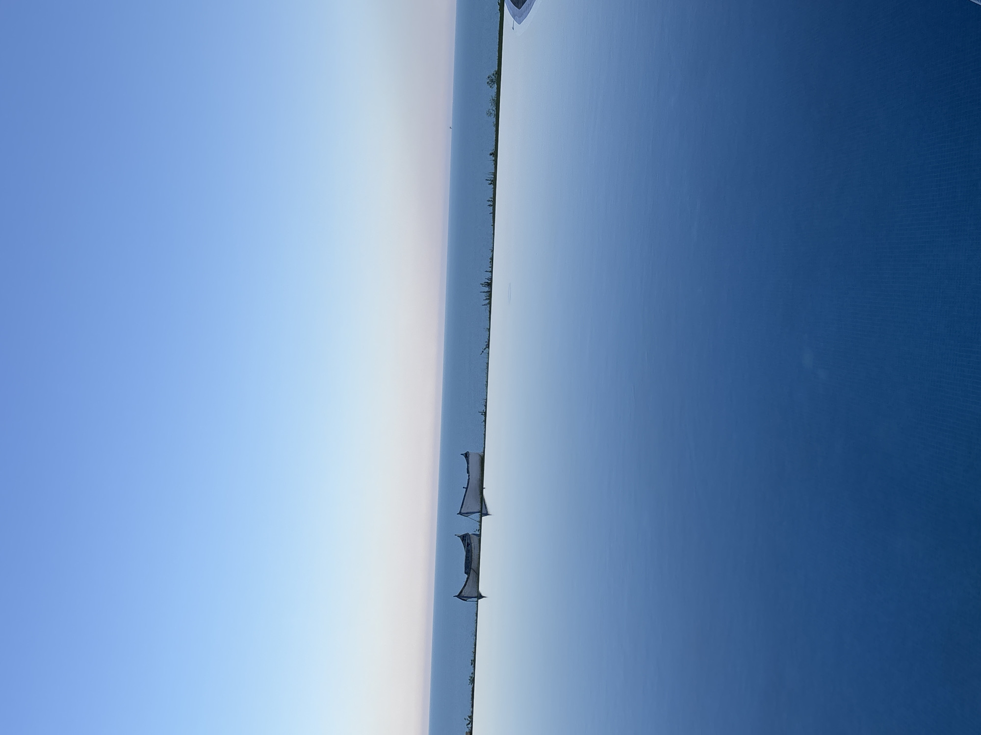 The image depicts an infinity pool overlooking the Black Sea in Topola, Bulgaria. Two small, white, wooden boats are positioned along the edge of the pool, on a strip of green vegetation separating the pool from the sea. The sky is clear and blue, transitioning to a lighter hue near the horizon. The water in the infinity pool is calm and reflects the sky and the boats. The scene conveys a sense of tranquility and scenic beauty.