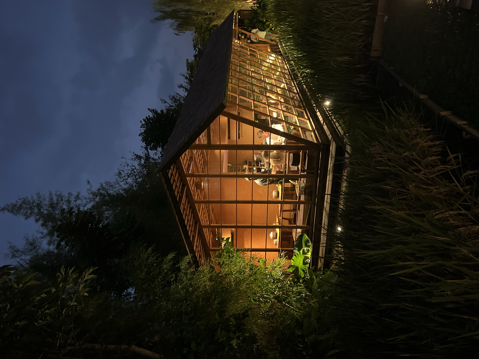 This night scene captures a modern, glass-paneled wooden cabin nestled within lush tropical vegetation. The cabin, featuring a distinctive A-frame-like roof and extensive windows, is brightly illuminated from within, casting a warm glow onto its immediate surroundings. Inside, several individuals are visible: one person stands or leans over a counter or table, while another is seated, suggesting an ongoing activity such as dining, working, or socializing. The interior is furnished with tables and chairs.
The cabin is enveloped by tall, dark green grasses or reeds, which may be part of rice paddies, and further back, darker silhouettes of trees or bamboo are visible against the night sky. Small, subtle spotlights on the ground illuminate sections of the surrounding foliage. The sky overhead is dark and cloudy, indicating either late dusk or nighttime. The contrast between the illuminated, cozy interior and the dark, natural exterior creates a tranquil and inviting atmosphere. No text is visible in the image. The setting, characterized by its unique architecture and tropical greenery, aligns with the location context of Ubud District, Indonesia.