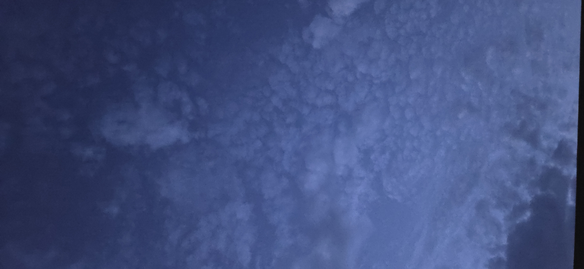 The image shows a dark blue sky filled with clouds. The clouds are varied in texture, with some appearing wispy and others more dense and textured. There is a horizontal dark band at the very bottom of the image, suggesting a foreground object such as a building or horizon line. The lighting suggests it is either dusk or dawn. The image does not depict any people, subjects, actions, or interactions. No specific location in Eeklo, Belgium can be determined from the provided image.