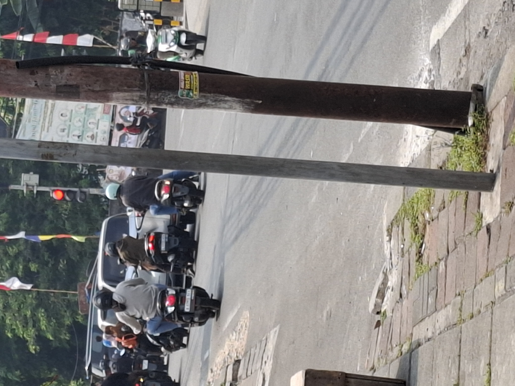 The image shows a street scene in West Jakarta, Indonesia, with several motorcycles and cars on a paved road. The foreground features a sidewalk made of bricks, with some grass growing through the cracks. A dark brown metal pole stands prominently on the right side of the image, with a black cable attached to it. A sticker with green and white lettering, possibly an advertisement or warning, is affixed to the pole.
In the background, several people are riding motorcycles. One person is wearing a light grey hoodie and blue jeans, another is wearing a dark jacket and dark pants, and a third person is wearing a light blue helmet and blue jeans. There are also cars, including a white vehicle, visible on the road. Traffic lights, with a red light illuminated, are present in the distance. Flags, red and white and red, white, and blue, are seen flying above the street. The overall impression is a busy urban environment during daylight hours.