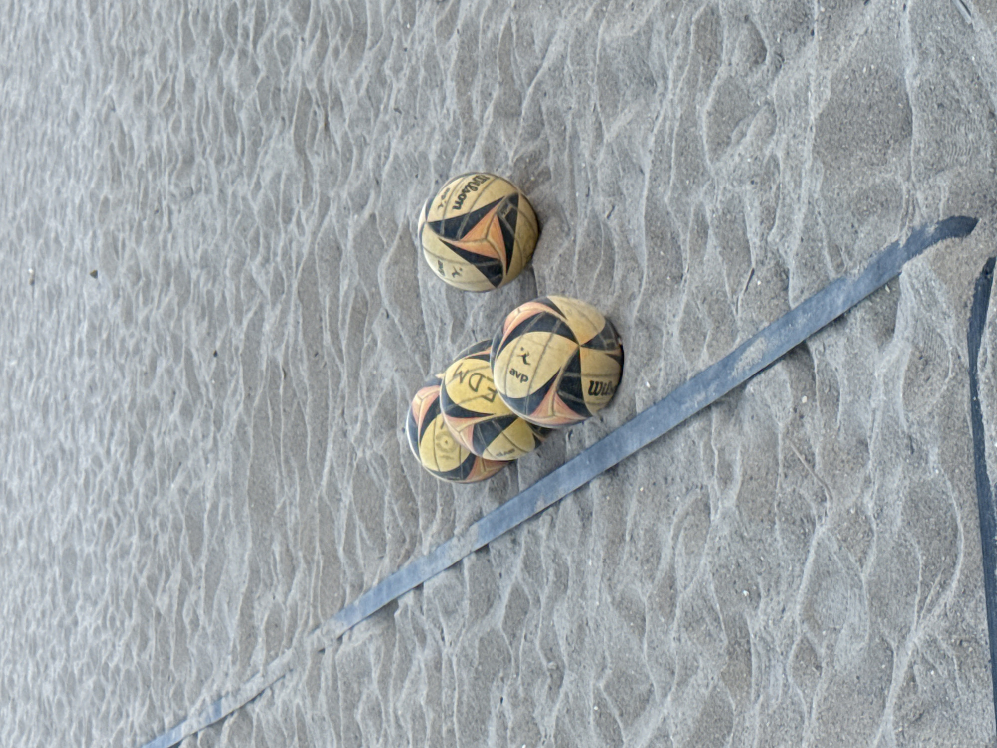 The image shows four volleyballs partially buried in the sand, resting against a dark blue volleyball net marker. The volleyballs are branded with "Wilson" and "AVP" logos. The sand is textured with windblown patterns. There are no people visible. The location is potentially a beach; however, the city of Barcelona cannot be verified from the image.