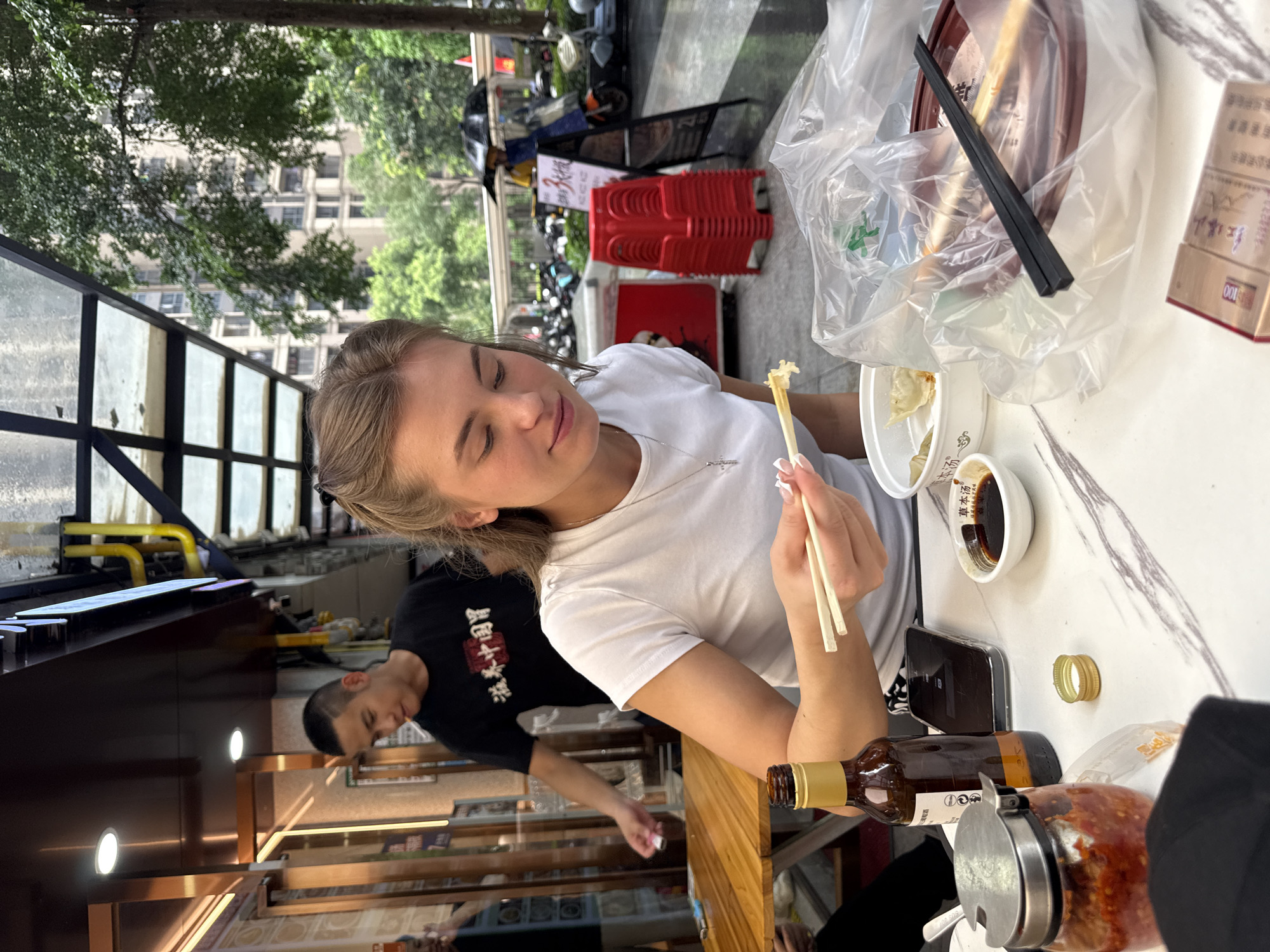 A young Caucasian woman with blonde hair tied back is seated at a marble-patterned table outdoors, eating with chopsticks from a white bowl. She is wearing a white t-shirt. In front of her is a small bowl of soy sauce, a bottle of amber liquid with a gold cap, a smartphone, a jar of red chili paste, and a package of cigarettes. A plastic bag contains a brown bowl and chopsticks. Behind her, a young Asian man wearing a black t-shirt with Chinese characters is standing and looking away. In the background, there are red plastic chairs stacked, a street with parked motorcycles, trees, and buildings. The scene appears to be in Haizhu District, China, indicated by the surrounding urban environment and the presence of Chinese text.