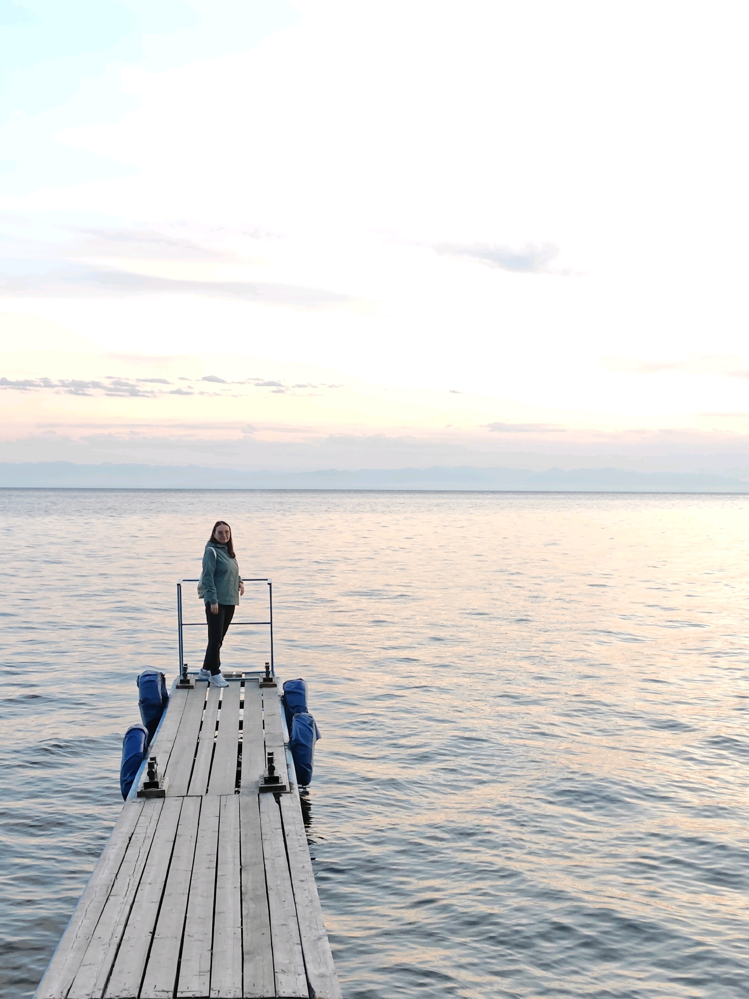 A young woman stands on a wooden dock extending into a large body of water, likely Lake Baikal, near Listvyanka, Russia. The sky above is a soft pastel blend of pink, orange, and blue, indicative of either sunrise or sunset. Distant, hazy mountains are visible on the horizon. The woman is wearing a green hooded jacket, dark pants, and white sneakers. She is looking towards the camera with a slight smile. The dock is constructed of weathered wooden planks and is supported by blue flotation devices on either side. The water is calm with gentle ripples reflecting the sky's colors.