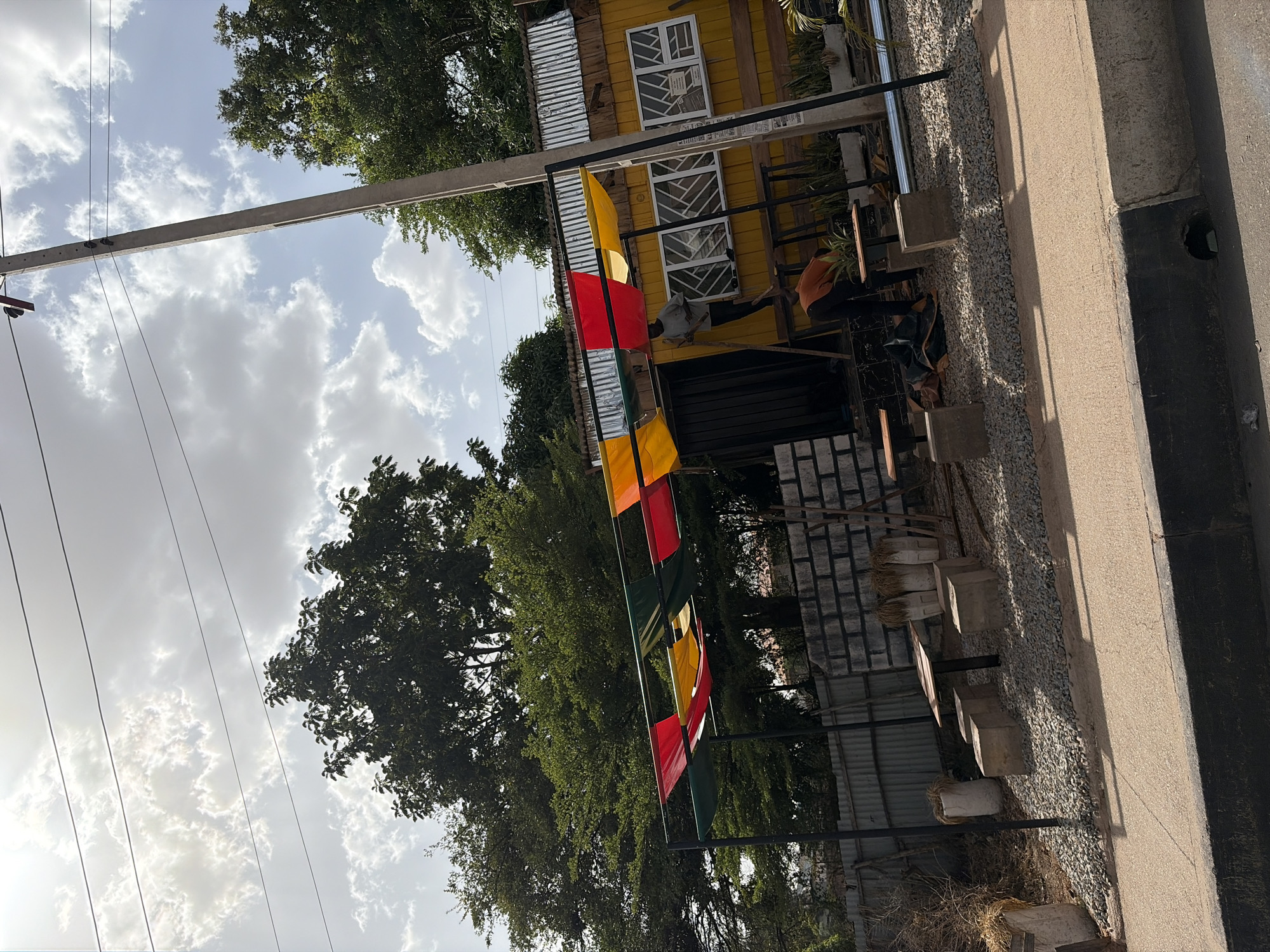 An outdoor scene in Jos, Nigeria, is depicted under a bright, cloudy sky during what appears to be daytime. Lush green trees frame the upper left and bottom left portions of the image. Power lines are visible stretching across the sky, anchored by a concrete utility pole on the left.
In the mid-ground, a row of colorful flags – primarily red, yellow, and green – are strung horizontally, adding a festive touch. Behind these flags, to the right, stands a vibrant yellow building or structure. It features white-framed windows, some of which have papers or flyers pasted on them (though text is illegible), and sections of corrugated metal siding.
Below the yellow building, the ground is covered in light-colored gravel. Several rough concrete blocks, possibly serving as seats or planters, are arranged on this gravel. Two individuals are present: one person wearing an orange shirt is seen sitting or working near some wooden planks in the foreground, while another person in a grey shirt stands closer to the yellow building.
To the far right, a concrete pathway or road runs parallel to the scene, with a dark drainage channel or opening next to it. The overall setting suggests a casual, possibly commercial or recreational, outdoor area.