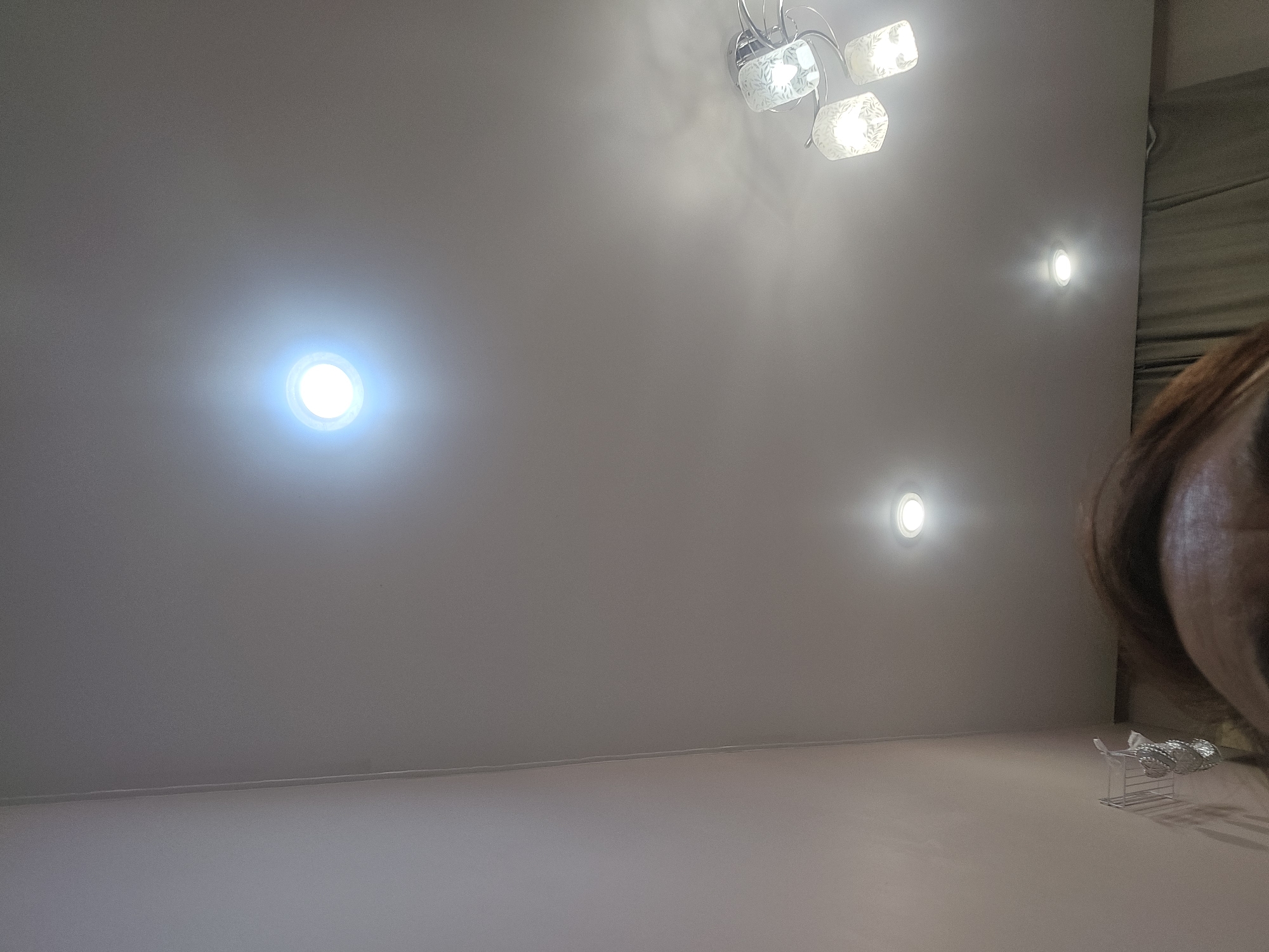 The image captures an indoor scene, likely a room, focused on the ceiling. A prominent feature is a bright, circular recessed light fixture emitting a cool white glow. To the right, a decorative ceiling light fixture with multiple glass shades, each with a subtle pattern, is visible. Further down and to the left, another smaller recessed light is also illuminated. The walls appear to be a light, neutral color. A portion of a person's head and shoulder is in the foreground, suggesting the photo was taken from a low angle, looking upwards. There is no indication of the city of Ulan-Ude, Russia, in the image.