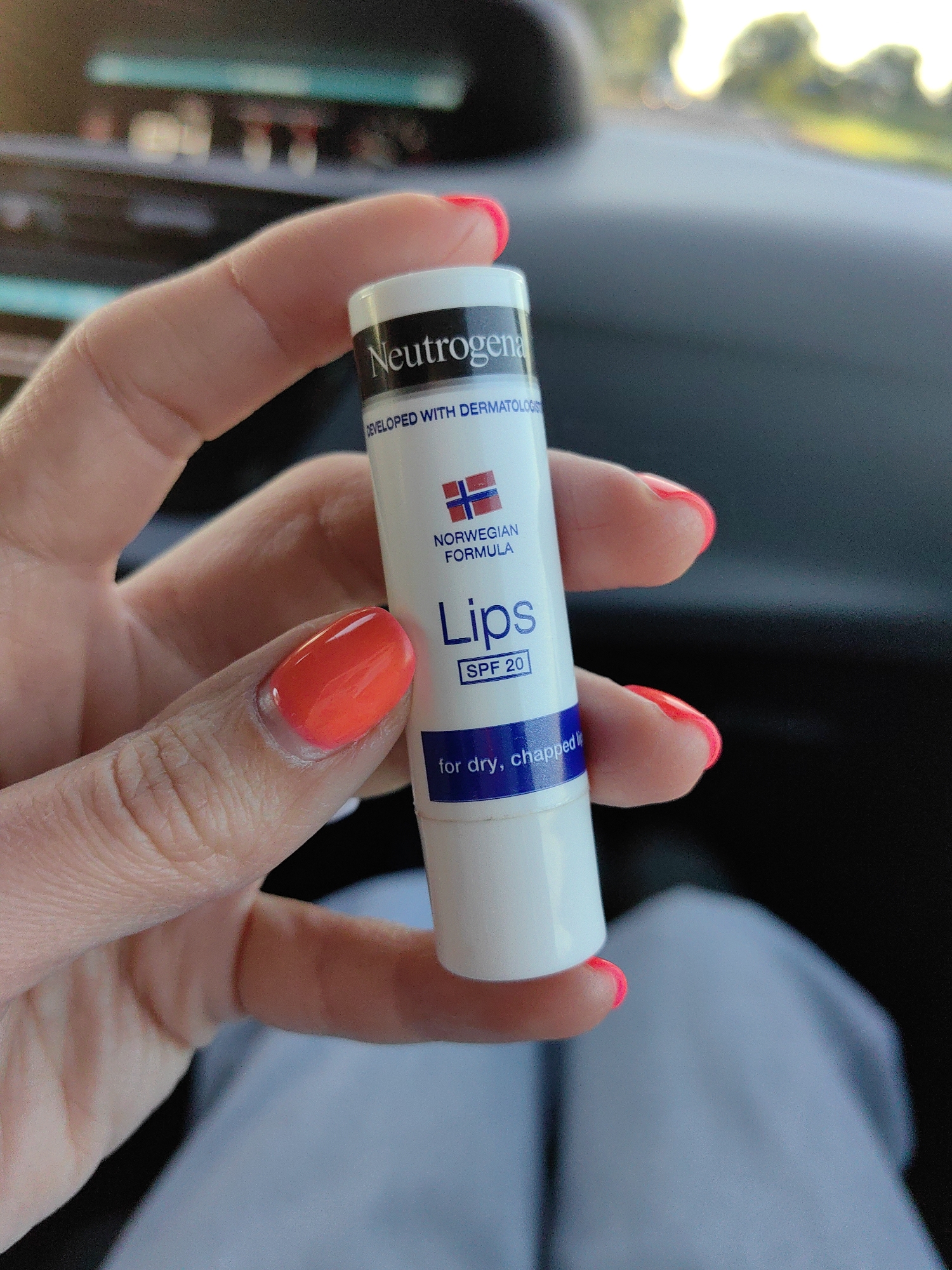 A person's hand, with bright orange nail polish, holds a Neutrogena lip balm stick. The lip balm tube is white with a black band at the top and text that reads "Neutrogena", "DEVELOPED WITH DERMATOLOGISTS", and "NORWEGIAN FORMULA". Below this, "Lips" is printed in large letters, followed by "SPF 20" in a blue box. Further down, the text "for dry, chapped li" is visible. The background is blurred and suggests an indoor environment, possibly a car, with hints of greenery and sunlight in the distance. The location in Дзяржынскі сельскі Савет, Belarus is not discernible from the image.