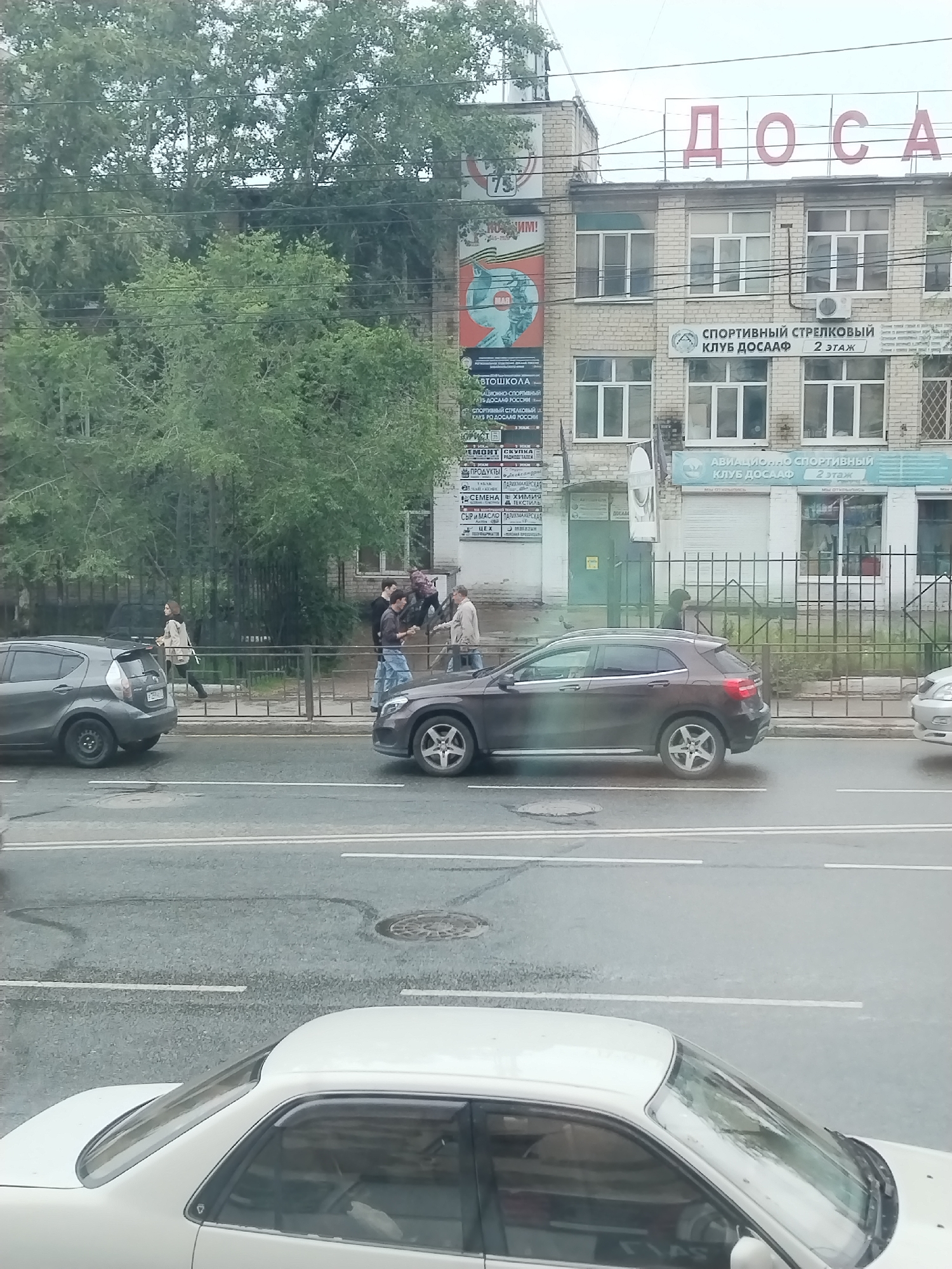 The image depicts a street scene in Chita, Russia, with a building in the background displaying Cyrillic signage, including "ДОСААФ". In the foreground, several cars are visible on the wet asphalt road. A dark gray hatchback is parked on the left side of the road, and a brown Mercedes-Benz SUV is positioned further down the street. A white sedan is partially visible at the bottom of the frame. On the sidewalk behind a metal fence, a group of four people are interacting. Two men in the center appear to be engaged in a discussion or argument, with one man in a light-colored jacket gesturing. Another man in a dark jacket is standing slightly behind them, and a fourth person in a light-colored jacket and dark pants is walking away from the group. Trees with green foliage are situated behind the fence, and overhead wires crisscross the scene. The overall lighting suggests an overcast day.