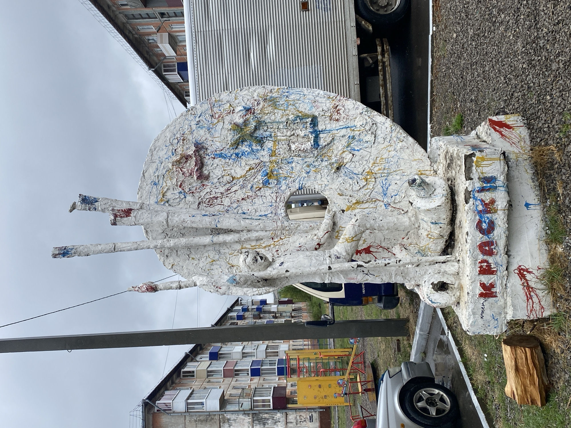 The image shows an outdoor sculpture in what appears to be a residential area of Usolye-Sibirskoye, Russia. The sculpture is primarily white with multi-colored paint splatters (red, blue, yellow) and features a disc-like shape with protruding extensions. It is positioned on a base covered in similar white material and paint. "KREACHOK" is written on the base in red paint, which is likely a misspelling of "Крючок", meaning "Hook" in Russian.
Behind the sculpture are residential buildings and a playground, suggesting a public space. A white commercial truck is partially visible behind the sculpture. The sky is overcast. A car and a wooden log are also visible in the foreground.