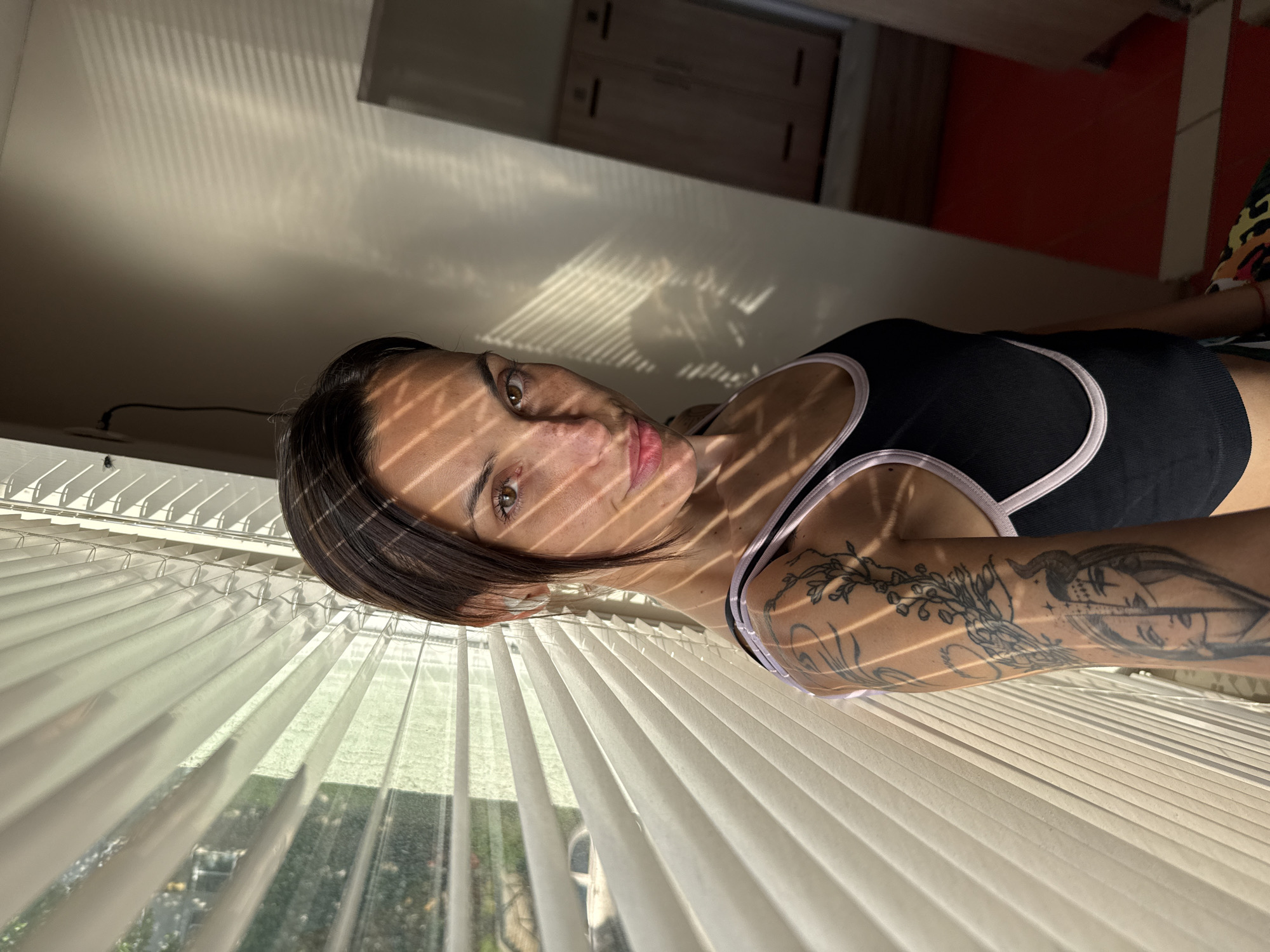 A woman is seen in a medium shot, lying down in front of white window blinds. The sunlight streams through the blinds, creating a striped pattern across her face. She has short brown hair and fair skin. She is wearing a black tank top with a light pink trim. A tattoo is visible on her left arm. Outside the window, greenery can be seen. The location is in a room with a red and white wall. The image provides no information on its location in Pleven, Bulgaria.