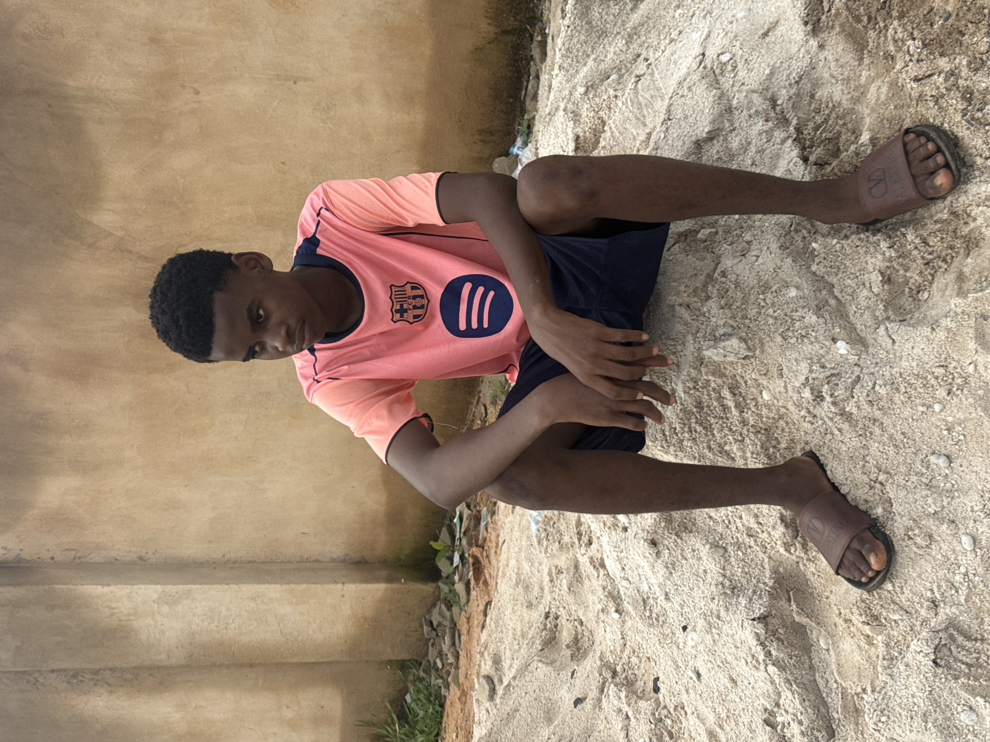 A young Black male sits with his legs extended on a sandy, uneven surface. He is wearing a pink t-shirt with navy blue trim and a navy blue logo on the chest, and navy blue shorts. On his feet, he wears brown slides with a prominent logo. His posture is relaxed, with one arm resting on his bent knee and the other arm extended. He is looking down towards the ground.
The setting appears to be outdoors, with a textured, beige-colored wall behind him on the left. The ground is covered in sand with scattered small white pebbles and some dark debris. Some sparse green vegetation is visible near the wall.
The lighting suggests it is daytime, with the sun casting some shadows. There is no visible text in the image other than the logo on the t-shirt and the slides. The overall impression is of a casual, unposed moment.
