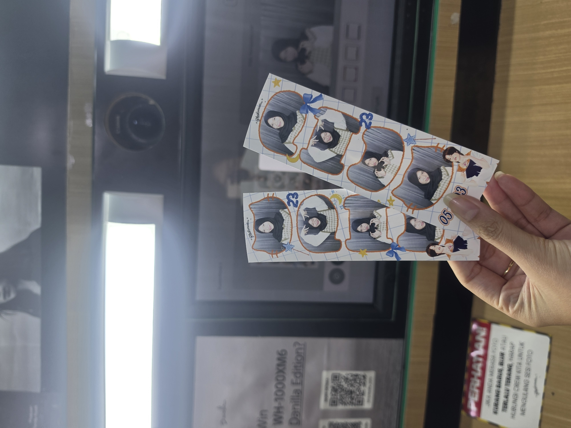 This media file captures a close-up view of a person's hand holding two photo strips from a photo booth. The photo strips feature stylized illustrations of two women, seemingly friends, posing in a playful manner. One strip has the numbers "23" and "053" printed on it, possibly indicating a date or a sequence number. The background reveals a photo booth, with its reflective surface showing another image of the women. A sign on the lower right corner in red and yellow reads "PERHATIAN" (Attention) followed by instructions in Indonesian, which seem to be related to photo quality. Below that, some text indicates "Win WH-1000XM6 Danilla Edition?" suggesting a promotional event. The scene appears to be indoors, possibly at a mall or entertainment venue in Medan, Indonesia, as suggested by the location context. The lighting is bright and artificial, typical of such locations. The overall mood is casual and fun, reflecting the enjoyment of taking commemorative photos.