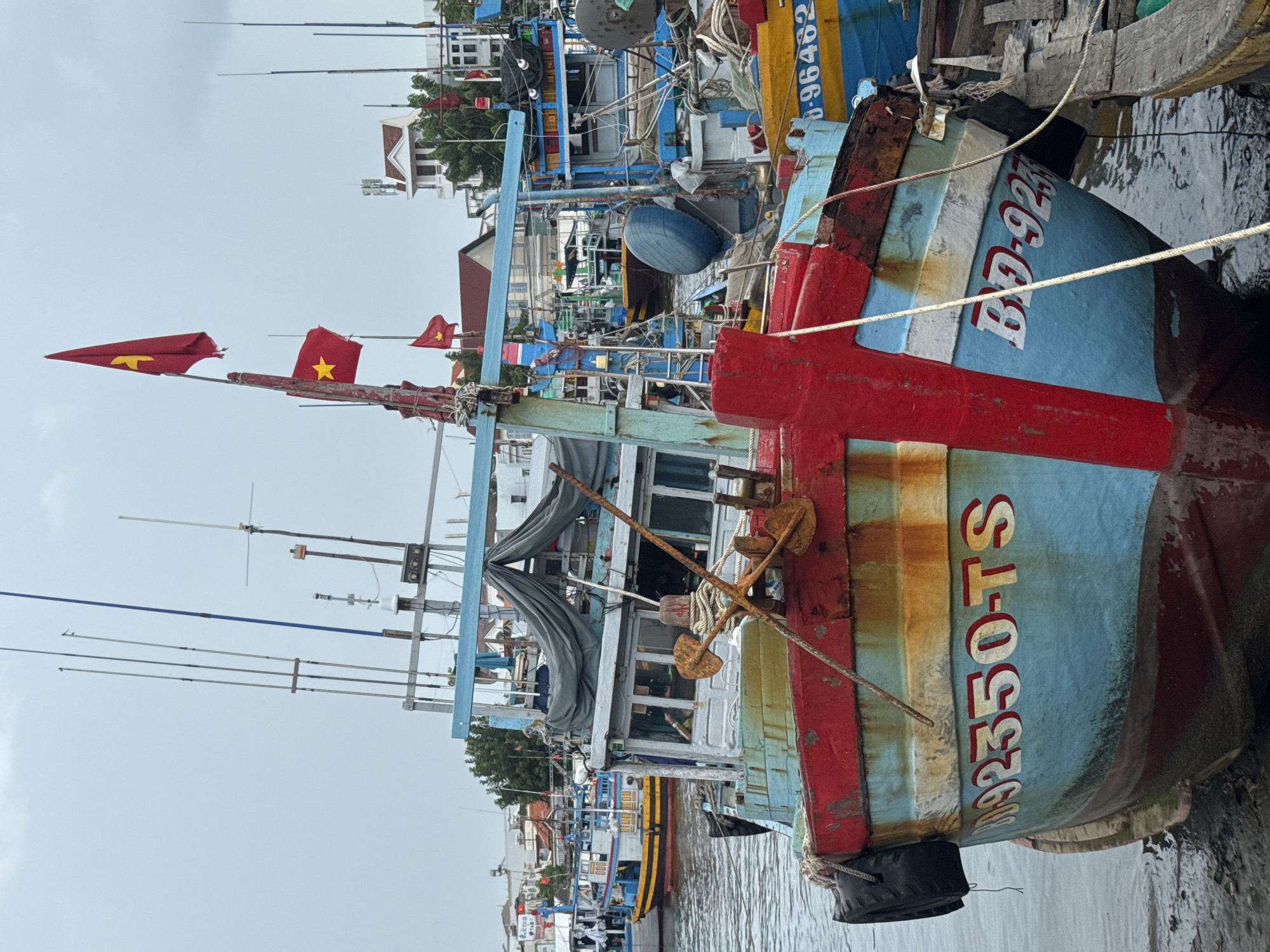 The image captures a close-up view of a light blue and red fishing boat docked in the city of Phan Thiết, Vietnam. The boat has a weathered appearance, with rust streaks visible on its hull. The boat's registration number, "BD-92350-TS," is painted in white with red outlines on its bow. Two Vietnamese flags, red with a yellow star, are prominently displayed on a pole at the front of the boat. Anchors and ropes are visible on deck. In the background, other fishing boats, buildings, and trees are seen, suggesting a bustling harbor. The sky is overcast. The water appears calm, and the boat is resting on a muddy shoreline.