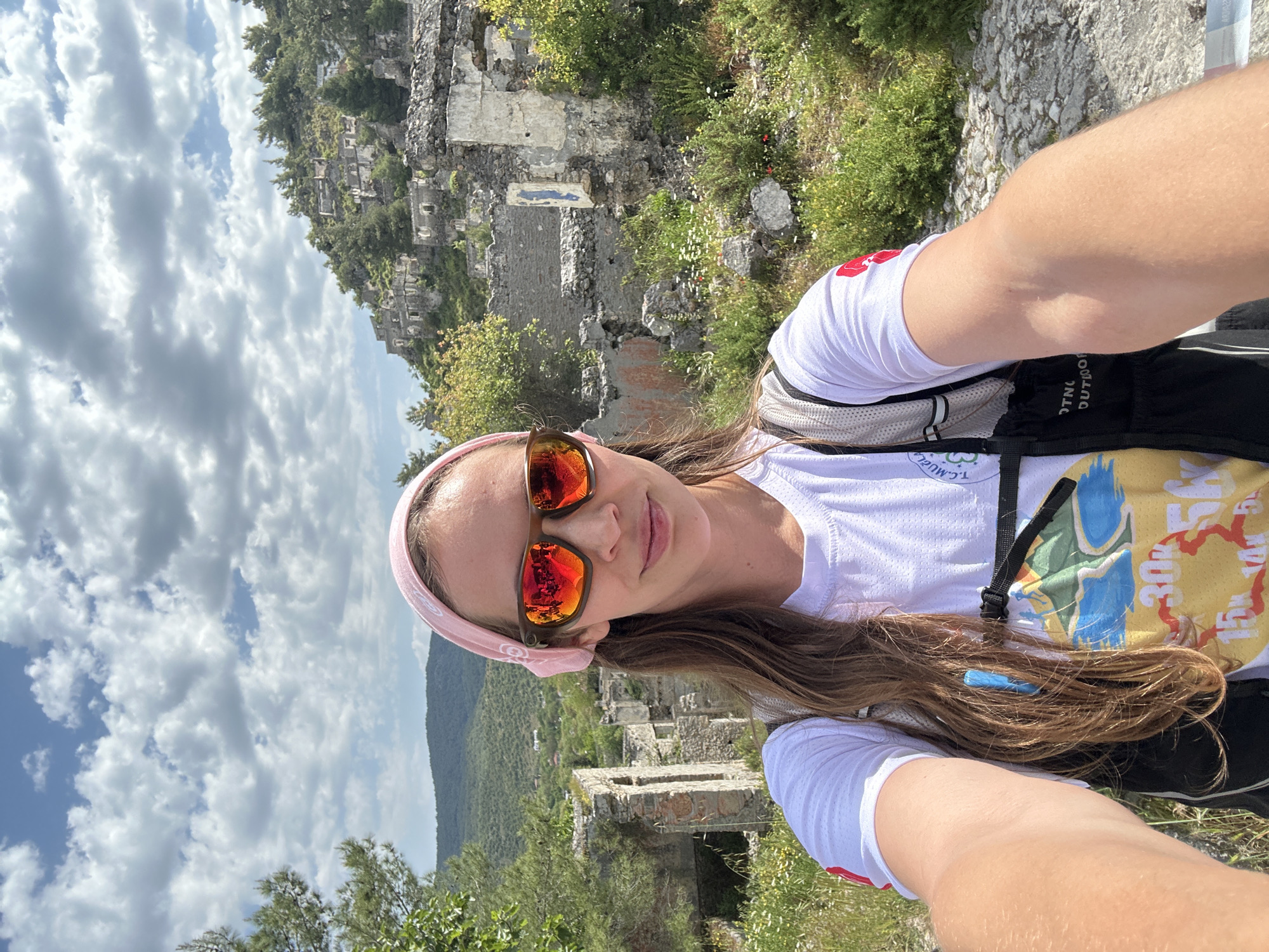 A woman is taking a selfie outdoors. She has light brown hair, sunglasses with orange lenses, and a pink headband. She's wearing a white athletic shirt and a black backpack with a colorful design. Behind her are stone ruins on a hillside, with greenery and a mountain range in the background. The sky is blue with fluffy clouds. The location is likely Kayaköy, near Fethiye, Turkey, judging by the style of the ruins and the landscape.