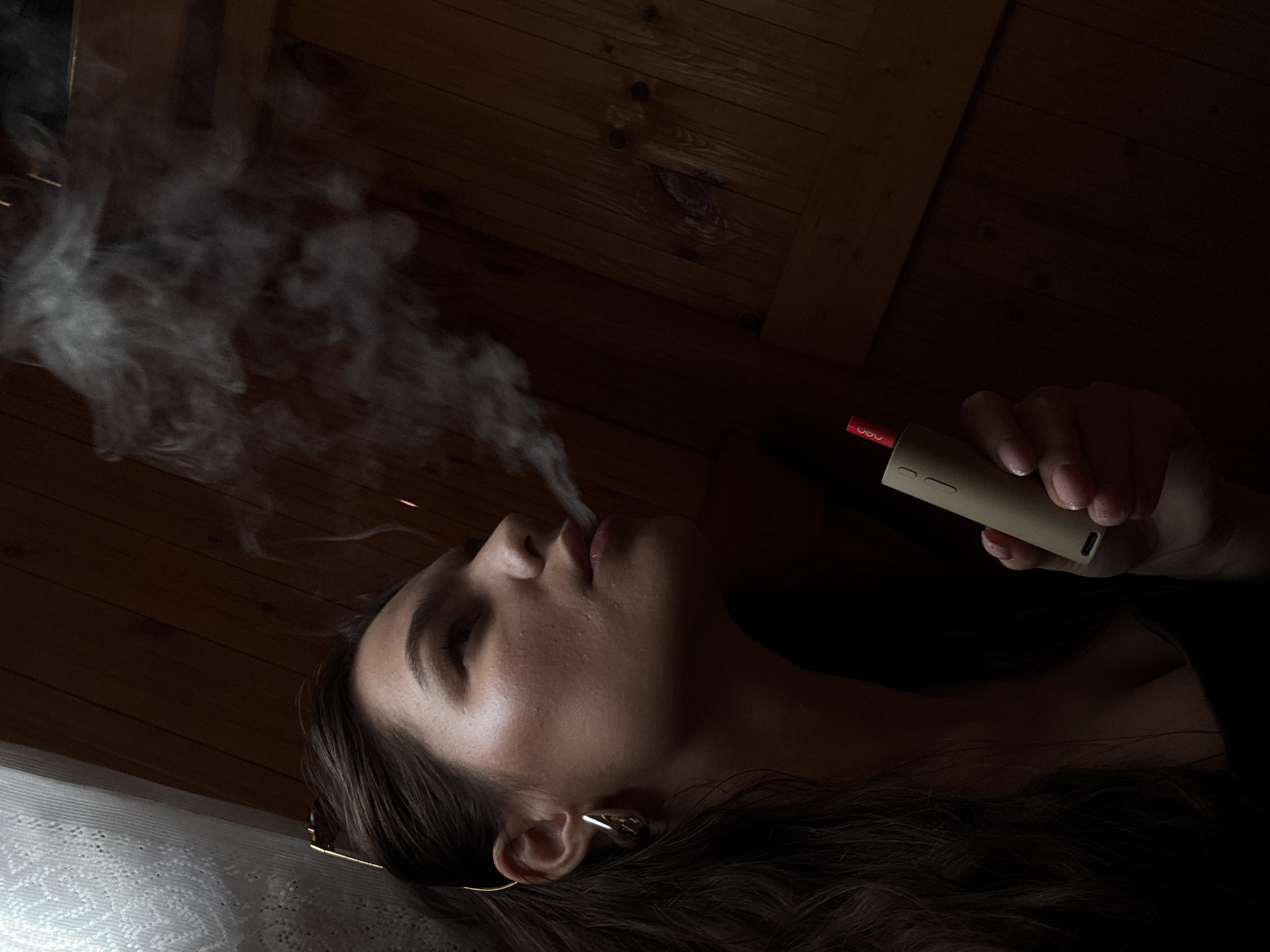 In a dimly lit interior space with wooden paneled walls, a young woman with dark hair is captured in a profile view, exhaling a plume of smoke. She is holding a beige vaping device with a red tip in her right hand. The woman is wearing a dark top and large, silver, teardrop-shaped earrings. Her gaze is directed upwards and to the right, away from the camera. The background consists of the textured wooden walls and a lacy white curtain to the left, suggesting an indoor setting. The location is stated to be in the city of Rusalevka, Russia.