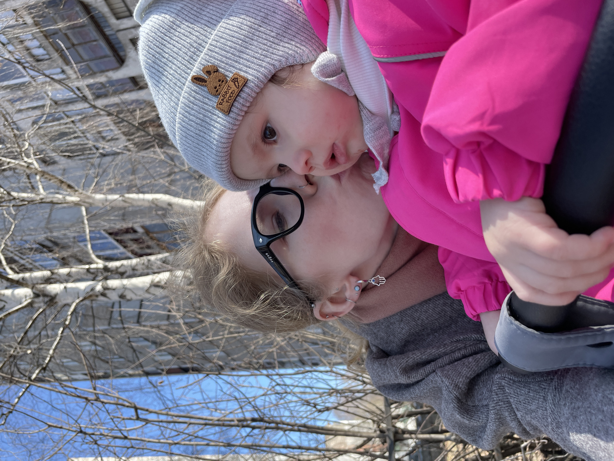 A medium shot depicts a woman holding a child against a backdrop of bare tree branches, a building facade, and a patch of blue sky. The woman wears black-framed glasses and a gray coat or jacket. The child is dressed in a bright pink jacket and a gray knit hat adorned with a patch. The child's footwear is gray and black. The photograph appears to be taken outdoors during daylight hours, and the environment suggests a park or urban setting. While it is not possible to verify the location as Izhevsk, Russia based on the visual information alone, the scene conveys an intimate moment of affection.