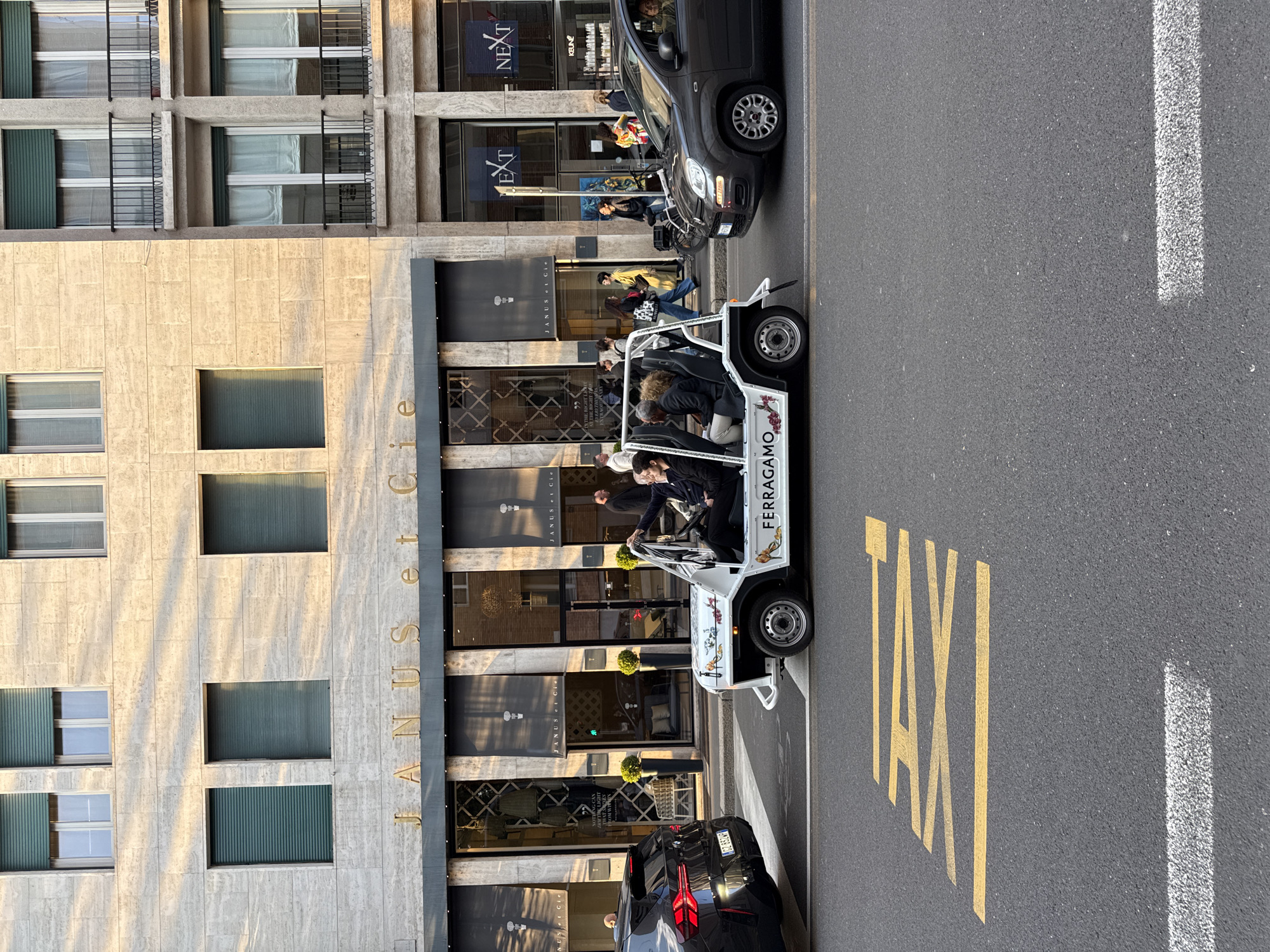 The image shows a street scene in Milan, Italy. A white, open-air taxi-like vehicle branded with "FERRAGAMO" is parked next to a curb in front of a building. The building has the name "JANUS et Cie" vertically displayed on its façade. Further down the street, another storefront is visible with "NEXT" displayed. Several people are walking on the sidewalk in front of the stores, and a black car is parked both in front and behind the Ferragamo vehicle. A yellow "TAXI" marking is painted on the asphalt, adjacent to a white lane line. The building's architecture includes multiple floors with windows and some with shutters. The overall scene conveys a busy city street in a commercial area.