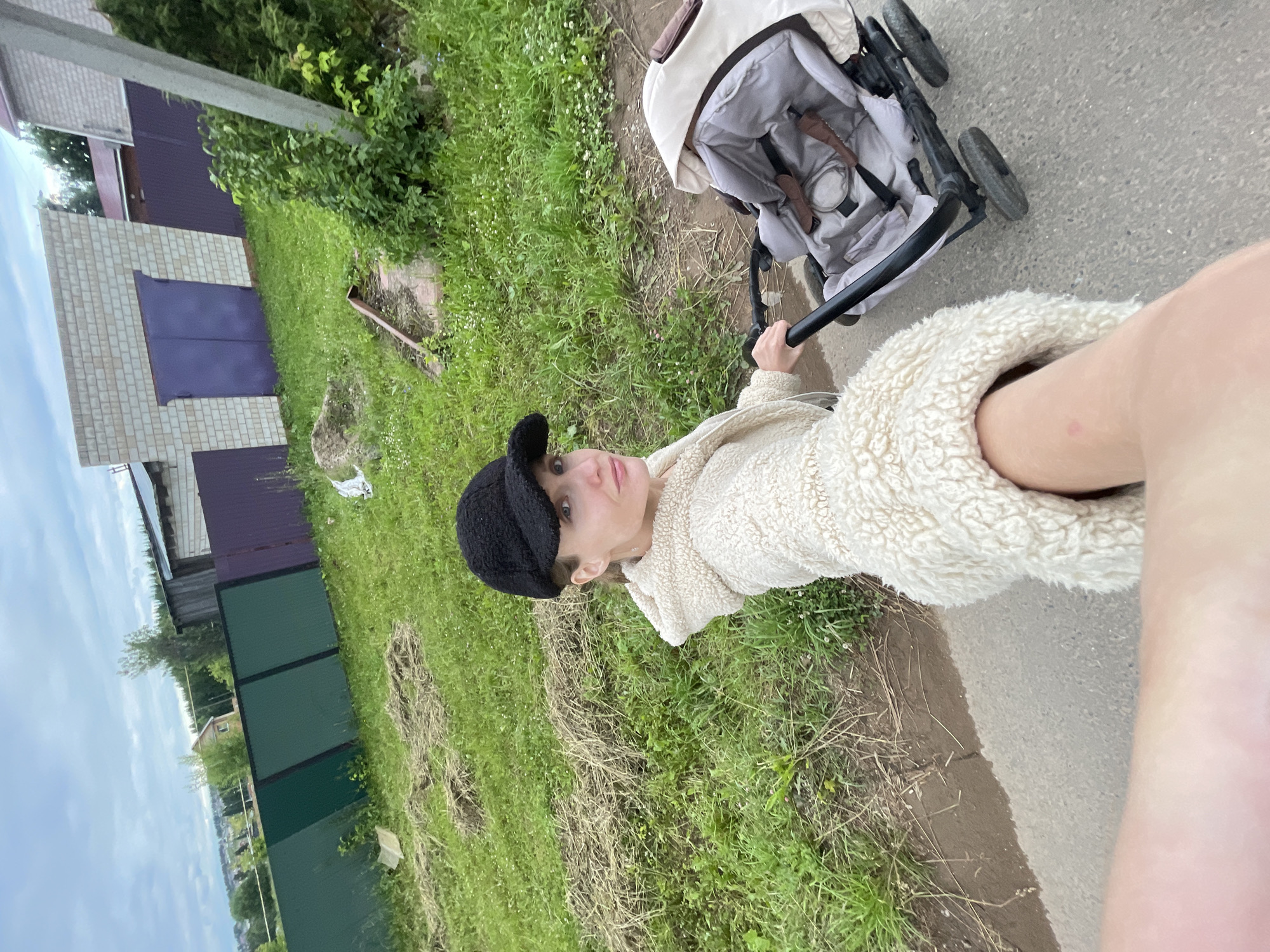 A person, wearing a cream-colored fluffy jacket and a black cap, pushes a stroller on a paved path next to a grassy area. The stroller is grey and beige. In the background, there is a building with a brick facade and dark purple doors, as well as a dark green fence. The sky is overcast, suggesting a cloudy day. The scene appears to be a residential area in the countryside, possibly during daylight hours. The person is looking at the camera with a neutral expression. There is no visible text in the image. The overall mood is casual and everyday.