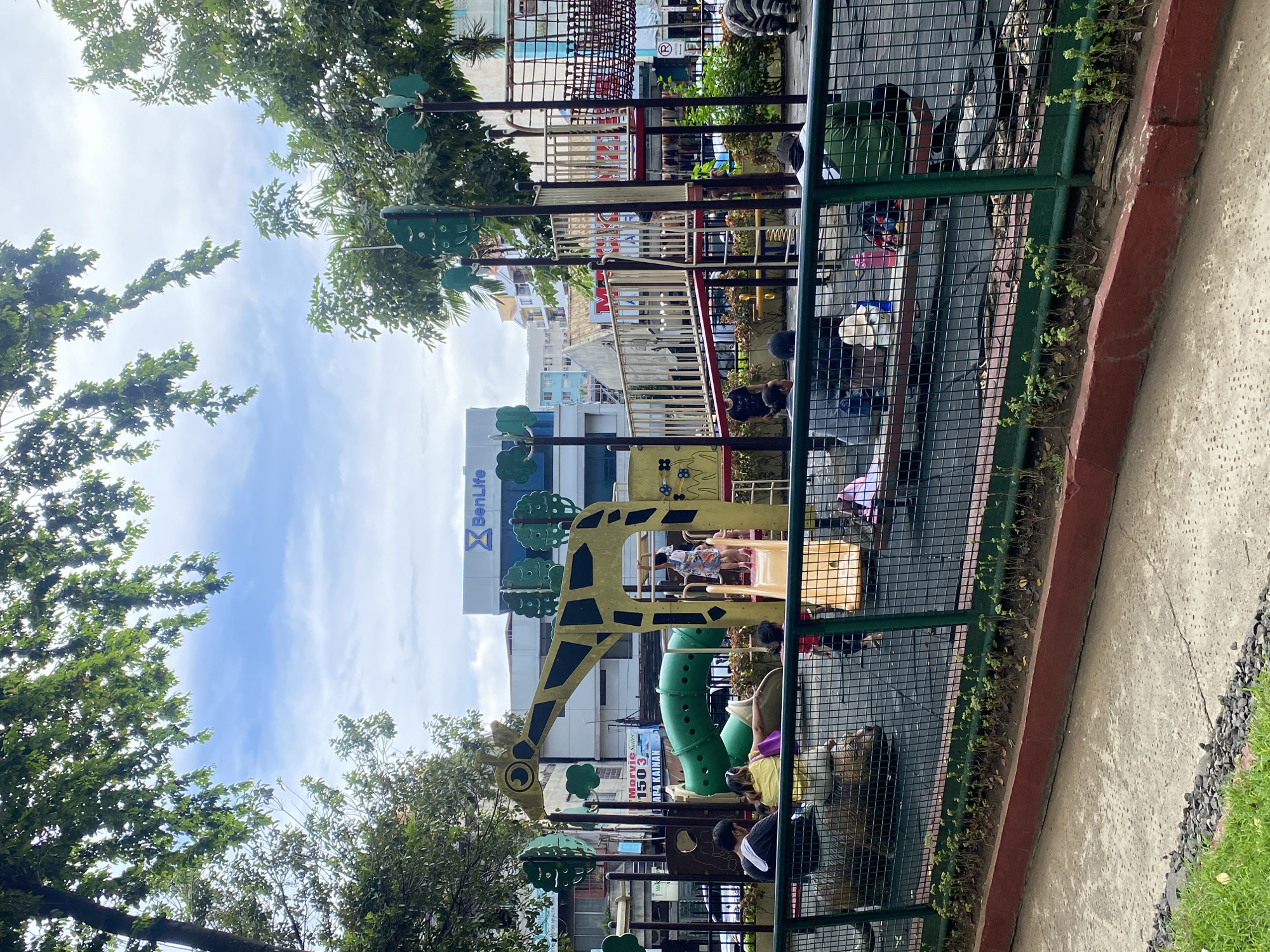 The image depicts a public playground in Davao City, Philippines, identified by the "BenLife" building in the background, a known insurance company with offices in the city. The playground features a colorful, giraffe-themed structure with slides and climbing elements. Several people, including adults and children, are present in the playground area, some engaged in play and others supervising. A green metal fence encloses the playground, with a row of small plants growing along the base of the fence. The ground surface within the playground appears to be a dark, cushioned material, possibly rubberized for safety. Beyond the playground, buildings and trees are visible, suggesting an urban setting. The sky is partly cloudy.