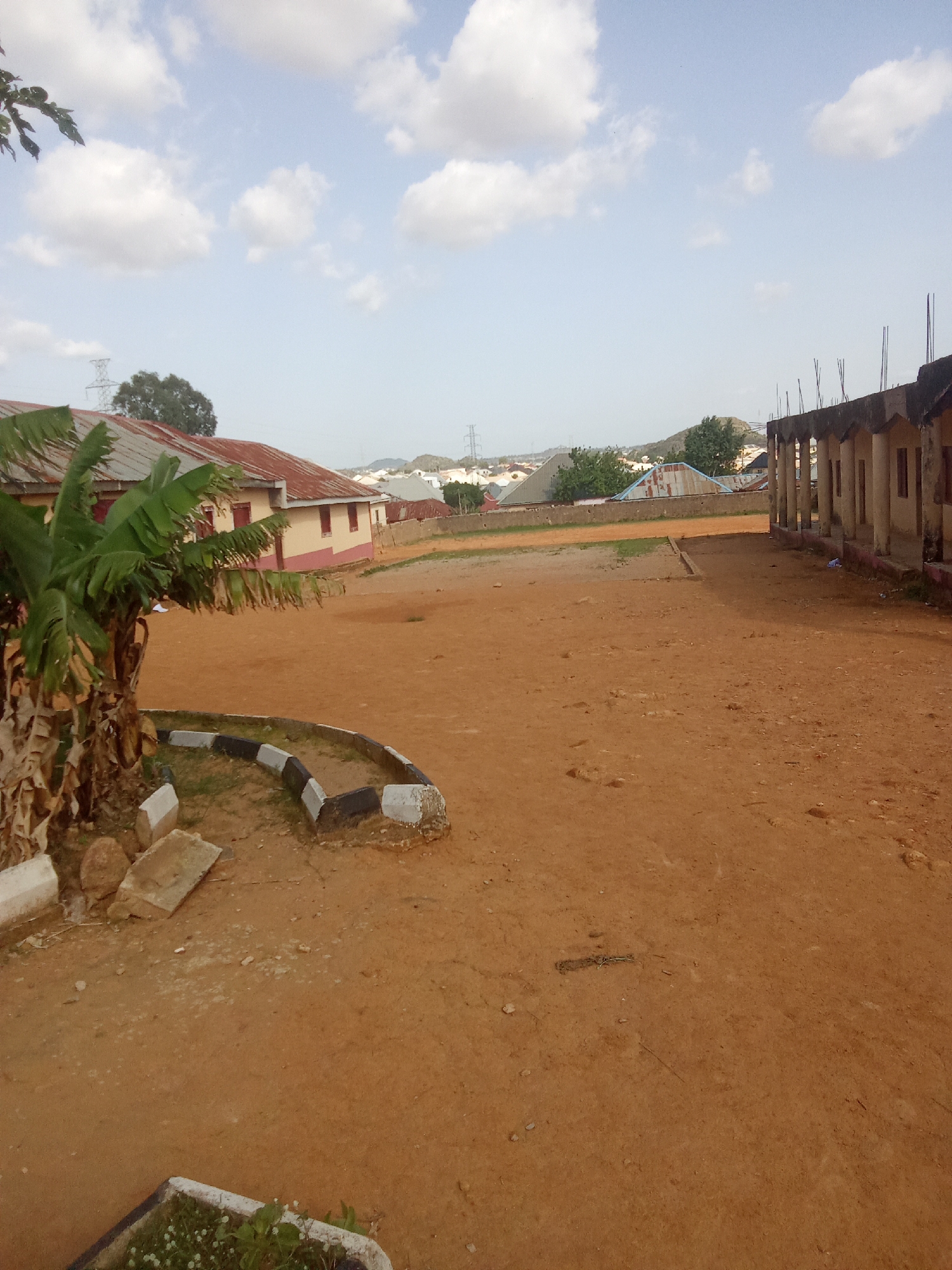 The image depicts an outdoor scene with a reddish-brown dirt ground and a pale blue sky with scattered white clouds. On the left side of the frame, there is a building with a corrugated red metal roof and pale yellow walls with reddish-brown trim. In front of this building, a banana tree with large green leaves stands prominently. To the right of the image, a partially constructed building with a concrete roof and support pillars is visible. The ground area appears to be a courtyard or open space. In the background, a hazy view of distant buildings and rolling hills can be seen. There are also some electrical towers visible in the far distance. The scene appears to be in a rural or semi-urban setting within the city of Barakin Akawo, Nigeria.