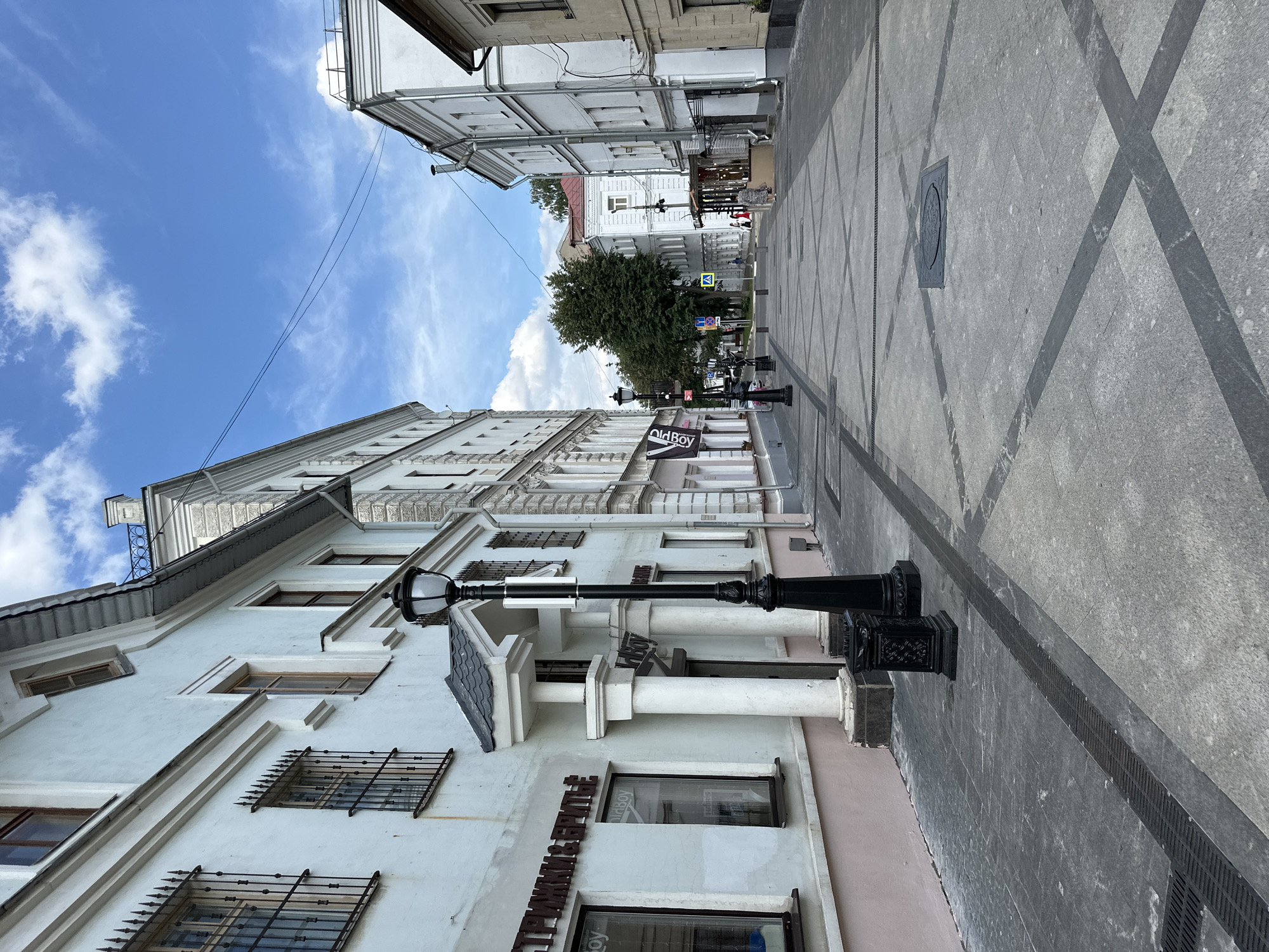 The image shows a street scene in Putyatino (Semivragovskaya S/A), Russia, on a partly cloudy day. The perspective is from a low angle, looking down a pedestrianized street lined with buildings.
On the left, a pale blue building features barred windows on the upper floors and a pink-painted lower section. The signage on the building is in Cyrillic and appears to advertise a barber shop or hair salon. A black lamppost with an ornate base stands prominently in the foreground. White columns support an overhang above an entrance to this building.
The street itself is paved with light gray stone tiles arranged in a pattern of intersecting lines, with darker gray diagonal stripes forming a grid. Grates are visible along the edges of the street.
On the right, a white building with multiple windows stretches down the street. Further down, other buildings of varying architectural styles and colors are visible, including a red-roofed building. A few people are present in the distance, walking along the street and near an outdoor seating area. Trees are visible further down the street, with green foliage. The sky above is blue with scattered white clouds.
