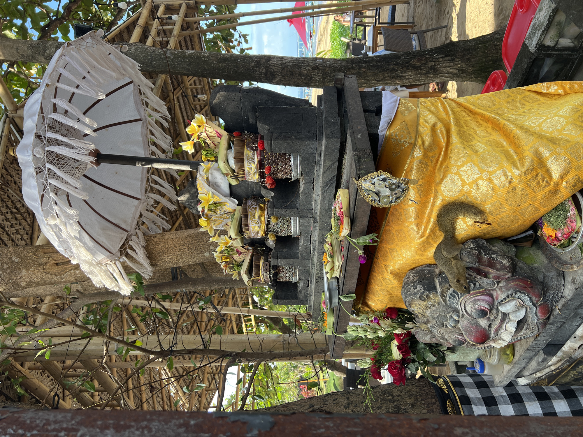 A Balinese Hindu shrine is set up outdoors, likely near a beach in Denpasar, Indonesia, given the presence of palm trees and beachgoers in the background. The shrine features a multi-tiered stone altar adorned with offerings of flowers, woven baskets filled with yellow flowers, and red pom-poms. A large white umbrella with fringe hangs over the altar. At the base of the shrine, a carved stone face, possibly a deity or demon, is visible, draped with a yellow cloth. A squirrel is perched on the stone face, interacting with the shrine. The surrounding environment includes trees, a thatched roof structure, and the sandy ground of a beach with tables and chairs visible in the distance.