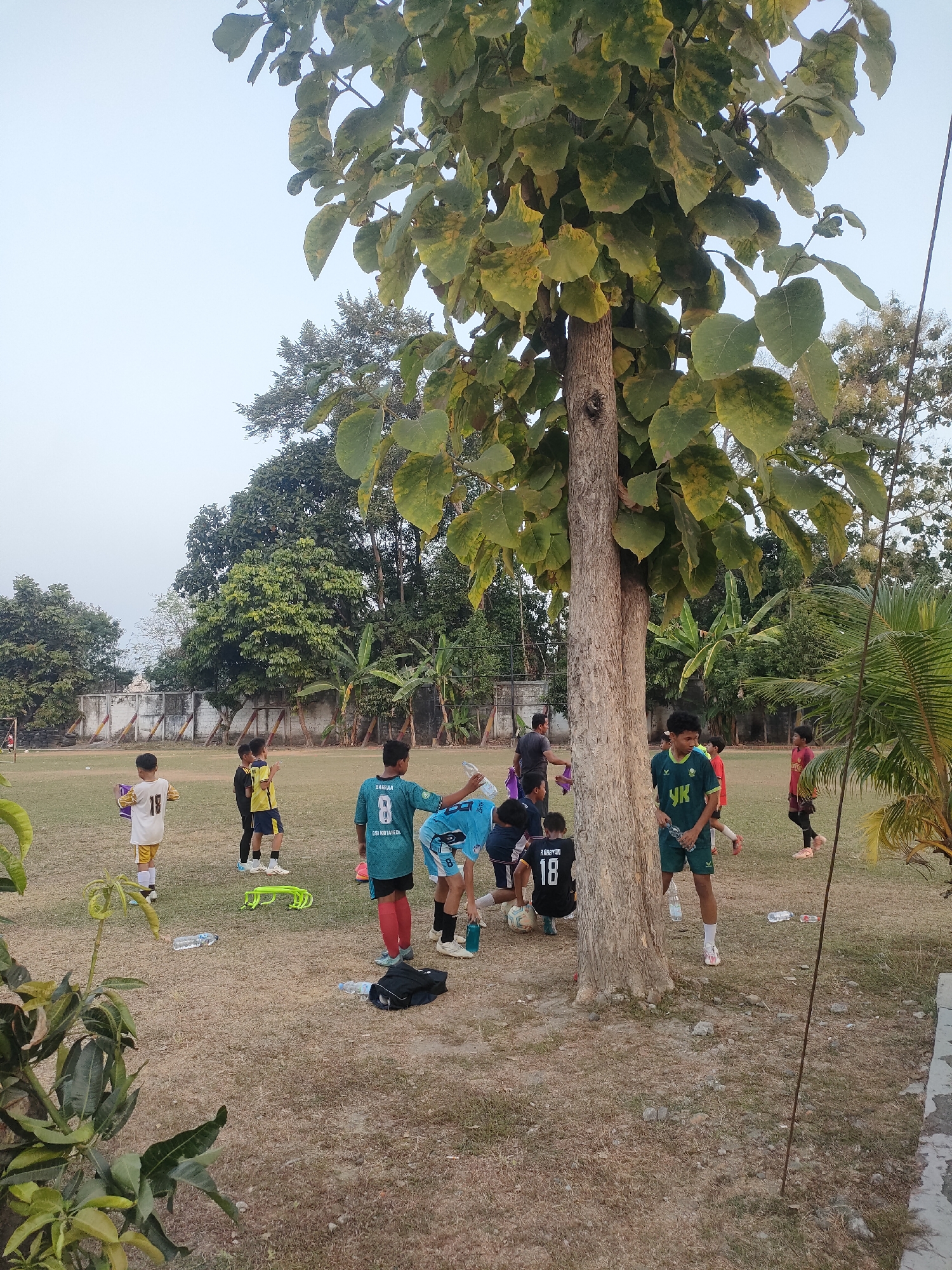 A group of young boys in soccer uniforms are gathered on a dry, grassy field near a large tree in Banguntapan, Indonesia. Some of the boys are standing and talking, while others are near soccer balls. The field appears to be part of a sports complex, with a fence and a building visible in the background. The sky is overcast.