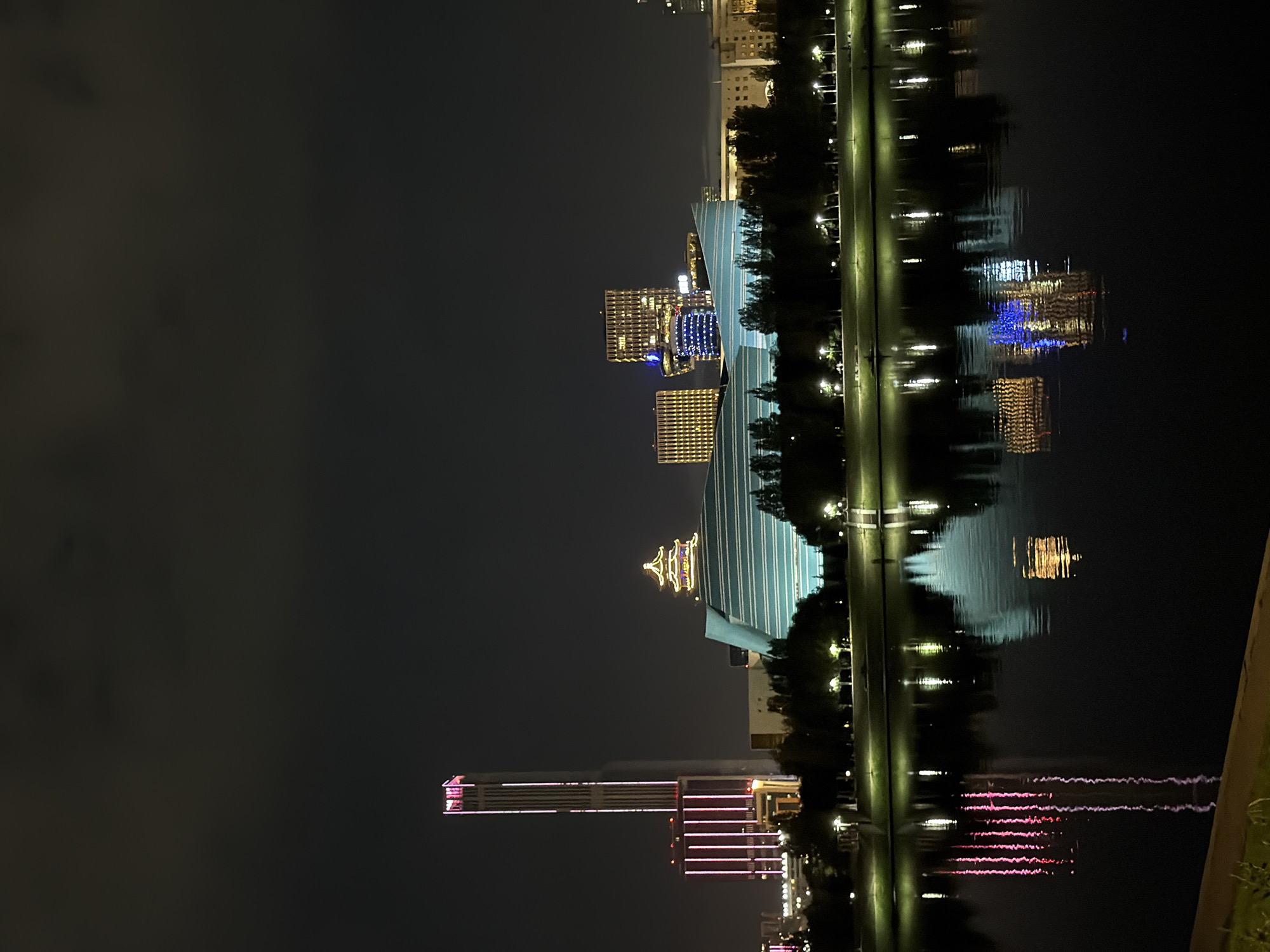 The image shows a nighttime view of the city of Astana, Kazakhstan, featuring modern architecture reflected in a body of water. In the background, a tall, slender skyscraper illuminated with pink vertical lights stands prominently. To its right, a distinctive building with a teal, curvilinear roof and a traditional-style, illuminated Asian pavilion on top is visible. Further to the right, several other modern buildings with illuminated windows and facades are present, including one with a blue and yellow patterned facade. A line of trees separates the buildings from the water. The water in the foreground reflects the lights of the buildings, creating a mirror image of the city skyline. The sky is dark and cloudy. The scene appears to be a park or waterfront area.
