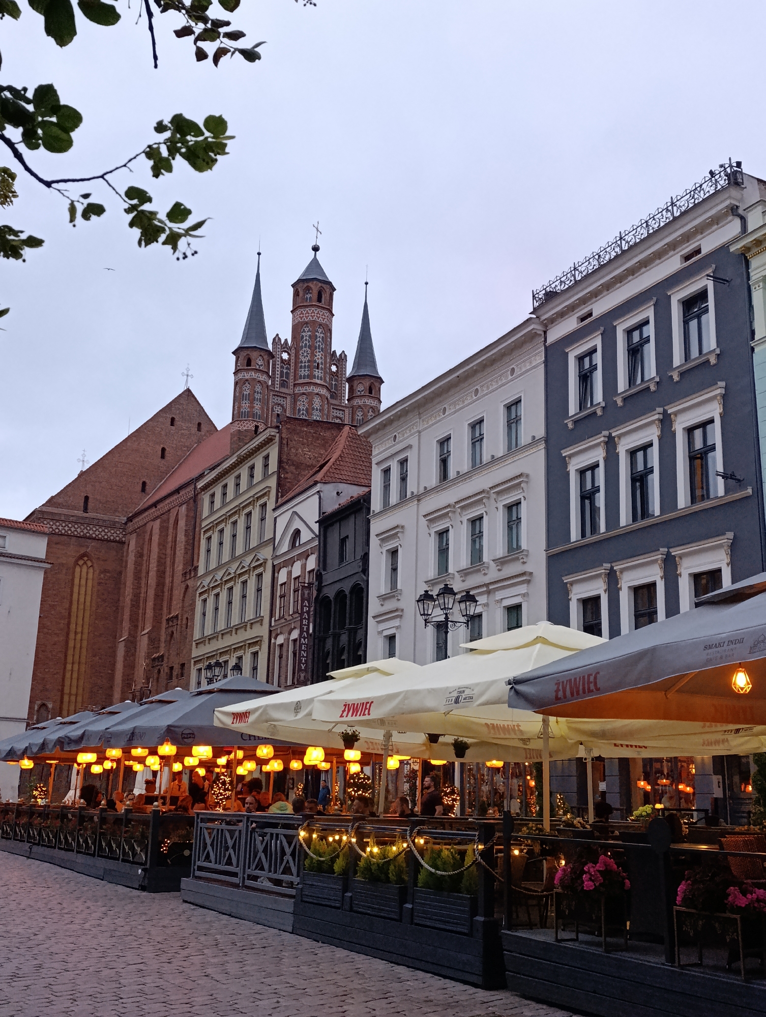 The image depicts a street scene in Toruń, Poland, likely during the early evening or overcast weather, given the artificial lighting present. In the background, a large, red brick Gothic church with multiple spires and intricate architectural details dominates the skyline. Adjacent to the church are several multi-story buildings with classical architectural styles, featuring rows of windows and ornate facades. In the foreground, a row of outdoor seating areas for restaurants or cafes is visible, covered by a series of large awnings. These awnings are illuminated by numerous small, warm-toned lights, creating a cozy ambiance. Patrons are seated at tables, some of whom are obscured by the awnings and the dim lighting. The ground appears to be paved with cobblestones. The overall scene suggests a European city with a rich architectural heritage and a vibrant outdoor dining culture.