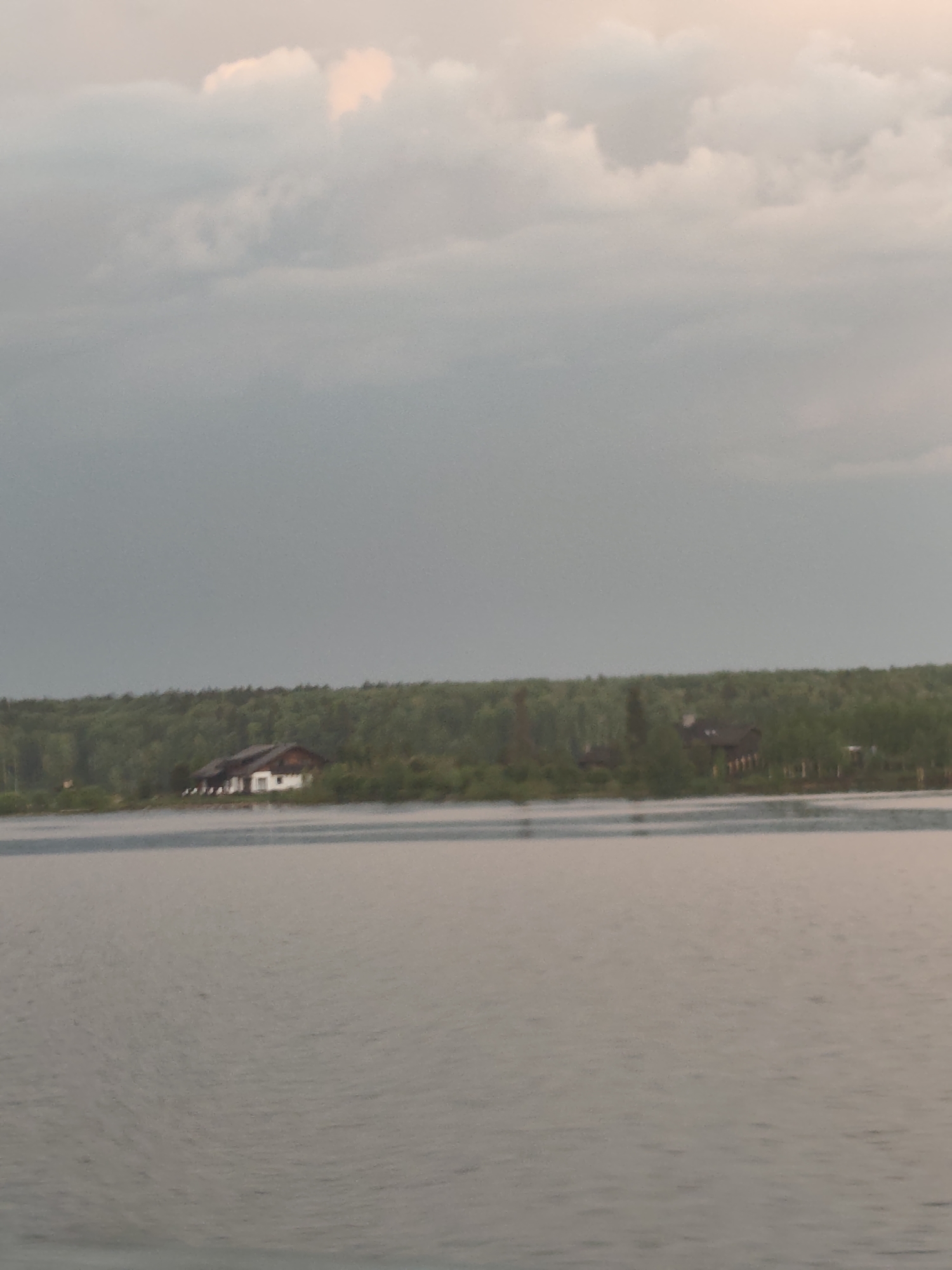 The image shows a body of water in the foreground, possibly a lake or river, with ripples visible on its surface. In the midground, there is a line of trees, likely a forest or shoreline. Several buildings are visible among the trees, including one on the left that appears to be a white house with a dark roof. The environment is overcast with gray clouds. The scene is Ust'-Shalashnaya, Russia.