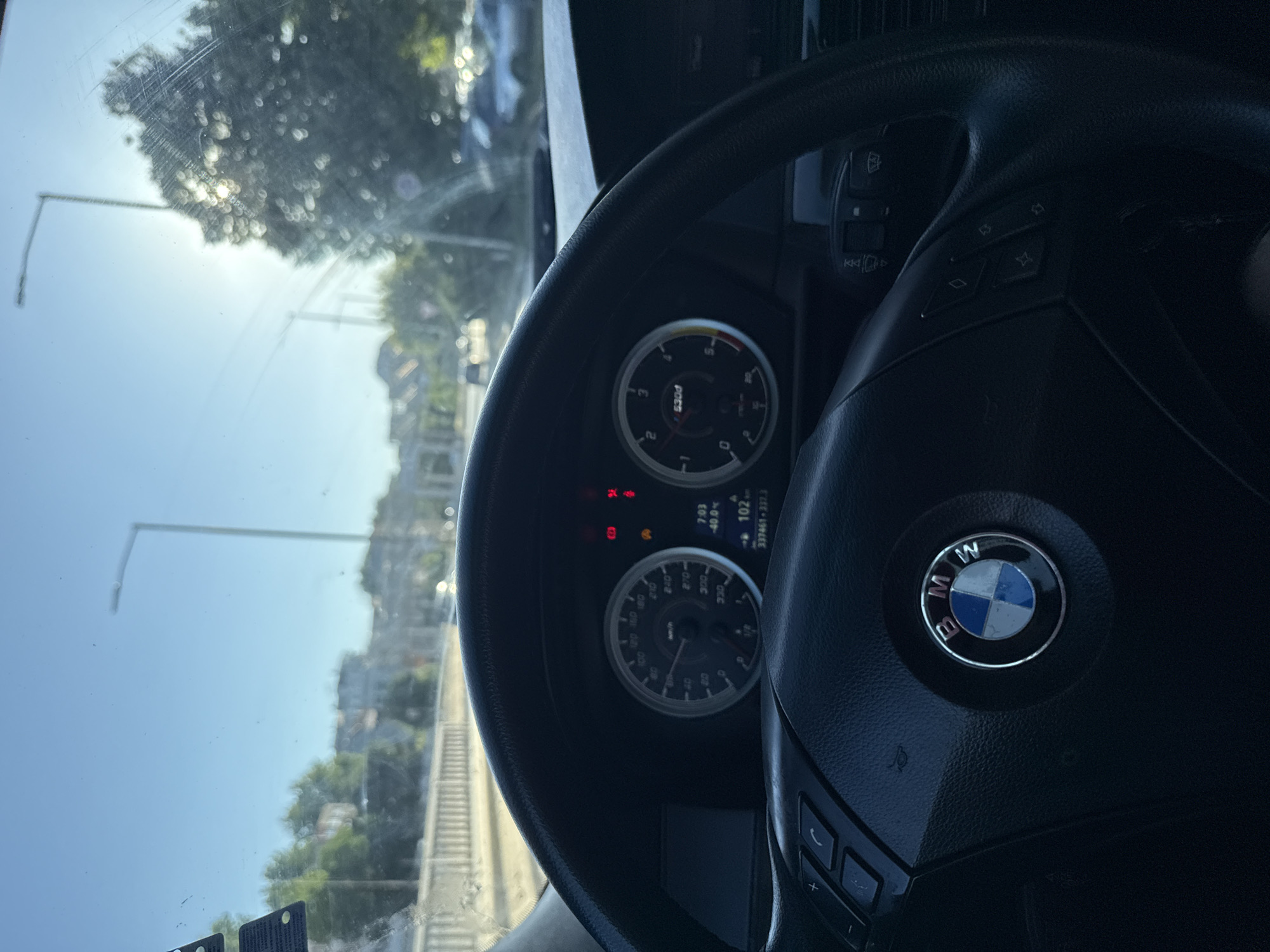 The image is taken from the interior of a BMW vehicle, looking out through the windshield. The sun is shining brightly, creating a glare and making some details difficult to discern. The dashboard and steering wheel are visible in the foreground. The steering wheel has the BMW logo in the center and controls on the left and right sides. The instrument cluster displays the speedometer, tachometer, and a digital screen showing time, temperature, and mileage. The exterior scene shows a street with traffic lights, streetlights, trees, and buildings. It appears to be daytime. The location is specified as the city of Varna, Bulgaria, though the specific street or area is not identifiable from the image alone.