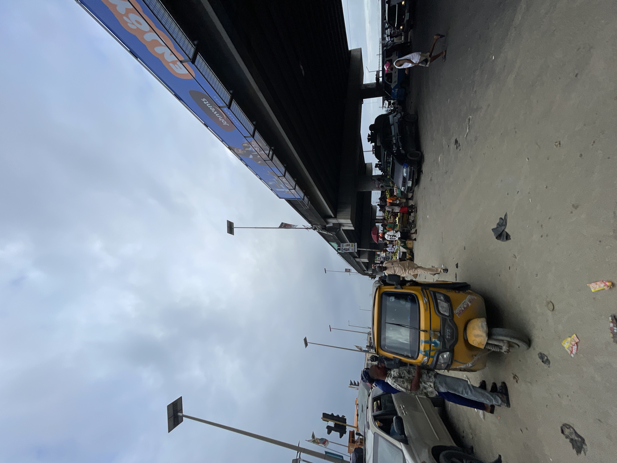 The image depicts a street scene in Lekki, Nigeria, characterized by a busy atmosphere beneath an elevated highway. In the foreground, a yellow auto-rickshaw is parked, with two men standing beside it. One man, wearing a patterned shirt and grey trousers, is leaning on the auto-rickshaw, while another man in a patterned shirt and dark trousers stands to his left. Further ahead, a silver car is visible.
The street is paved with a mixture of dirt and asphalt, with scattered litter visible on the ground, including several small packets and what appear to be discarded items of clothing. A market stall with fruits and vegetables is set up under the highway. Several people are browsing the market and walking around.
The elevated highway dominates the upper portion of the image, with a large blue billboard displaying text and images. Multiple streetlights with solar panels are positioned along the side of the road and beneath the highway. Traffic appears to be present, with several cars and another auto-rickshaw visible further down the road. The sky is overcast with a grey, cloudy appearance. The overall impression is one of urban congestion and commercial activity.