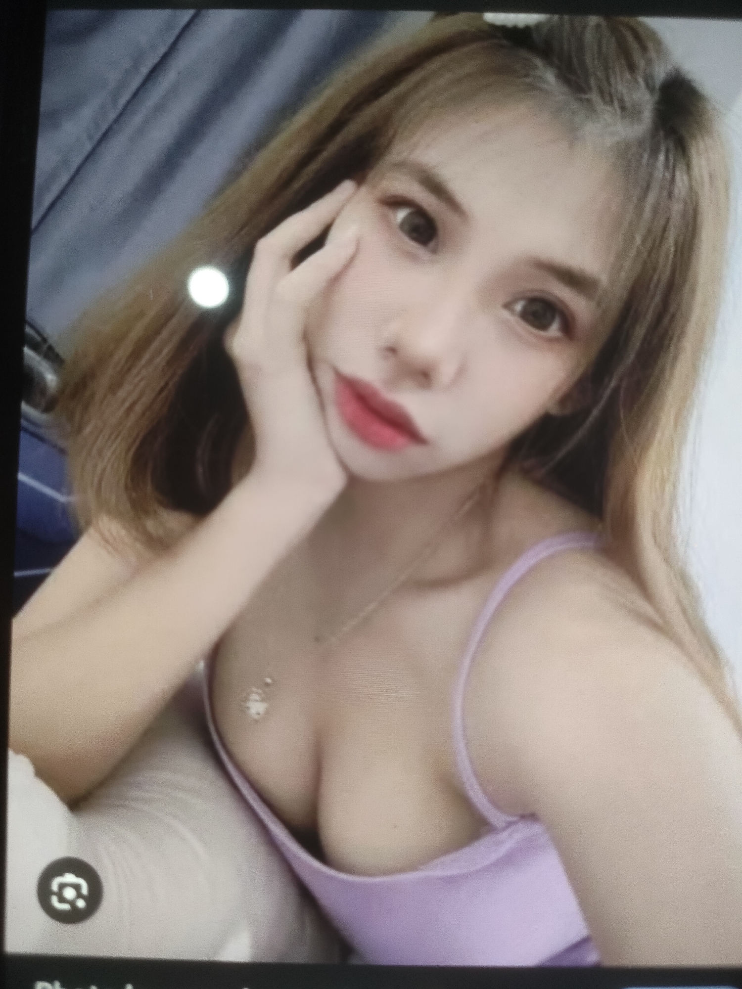 The image shows a close-up of a young woman with light brown hair and fair skin. She is wearing a light purple camisole. Her right hand is touching her cheek. The background appears to be a gray curtain. There is a small, white circular reflection on her hair. The image is slightly out of focus. No location in the city of Padang Dalom, Indonesia, is identifiable.