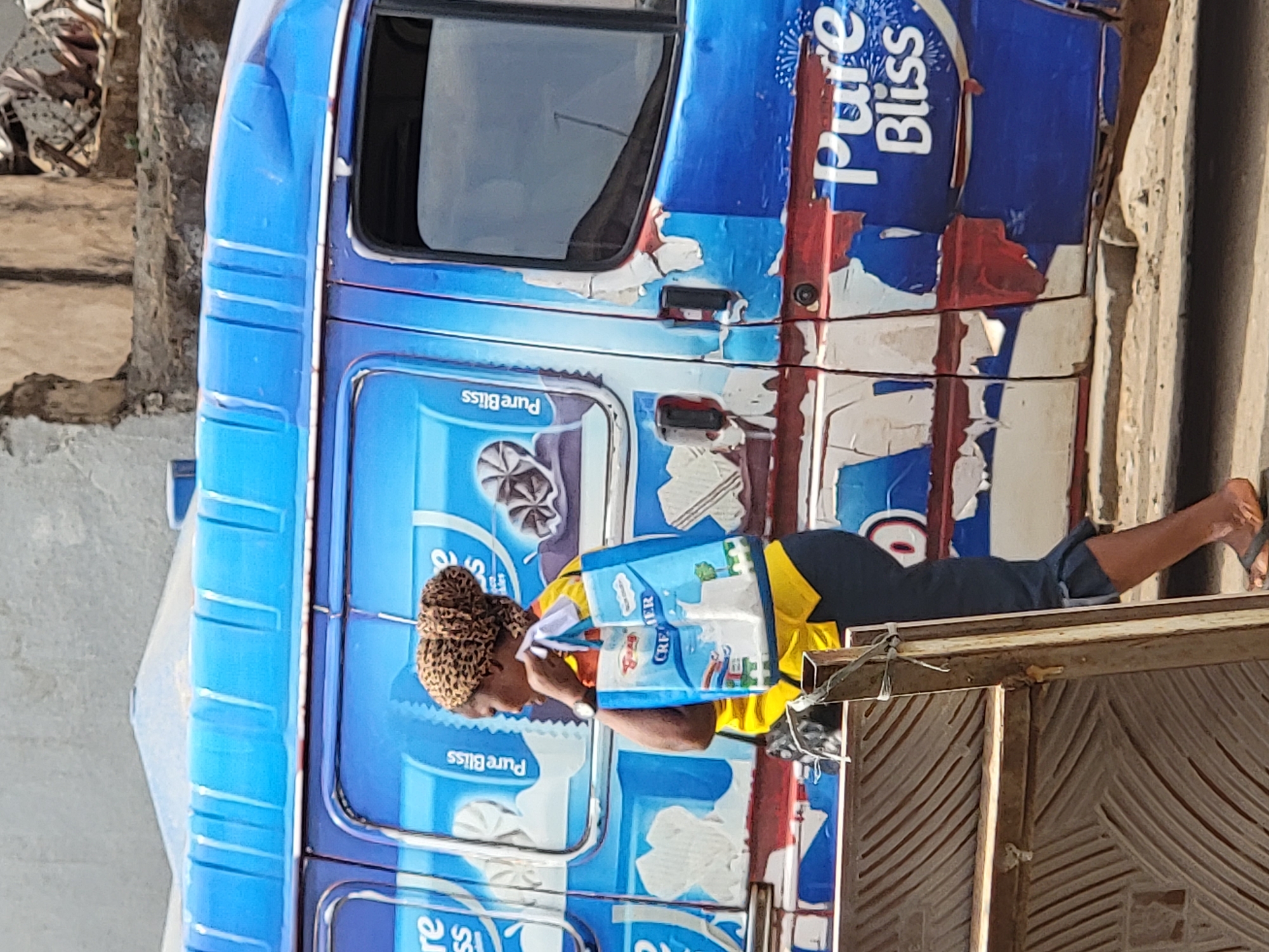 A woman is shown standing in front of a "Pure Bliss" brand truck in Maiburiji, Nigeria. She is wearing a yellow shirt, dark pants, and a leopard-print head wrap, and is carrying a "CREMER" branded bag. The truck is predominantly blue and white with faded and peeling paint. The setting appears to be outdoors in an urban environment, with a building with a textured door in the foreground and signs of street activity in the periphery.