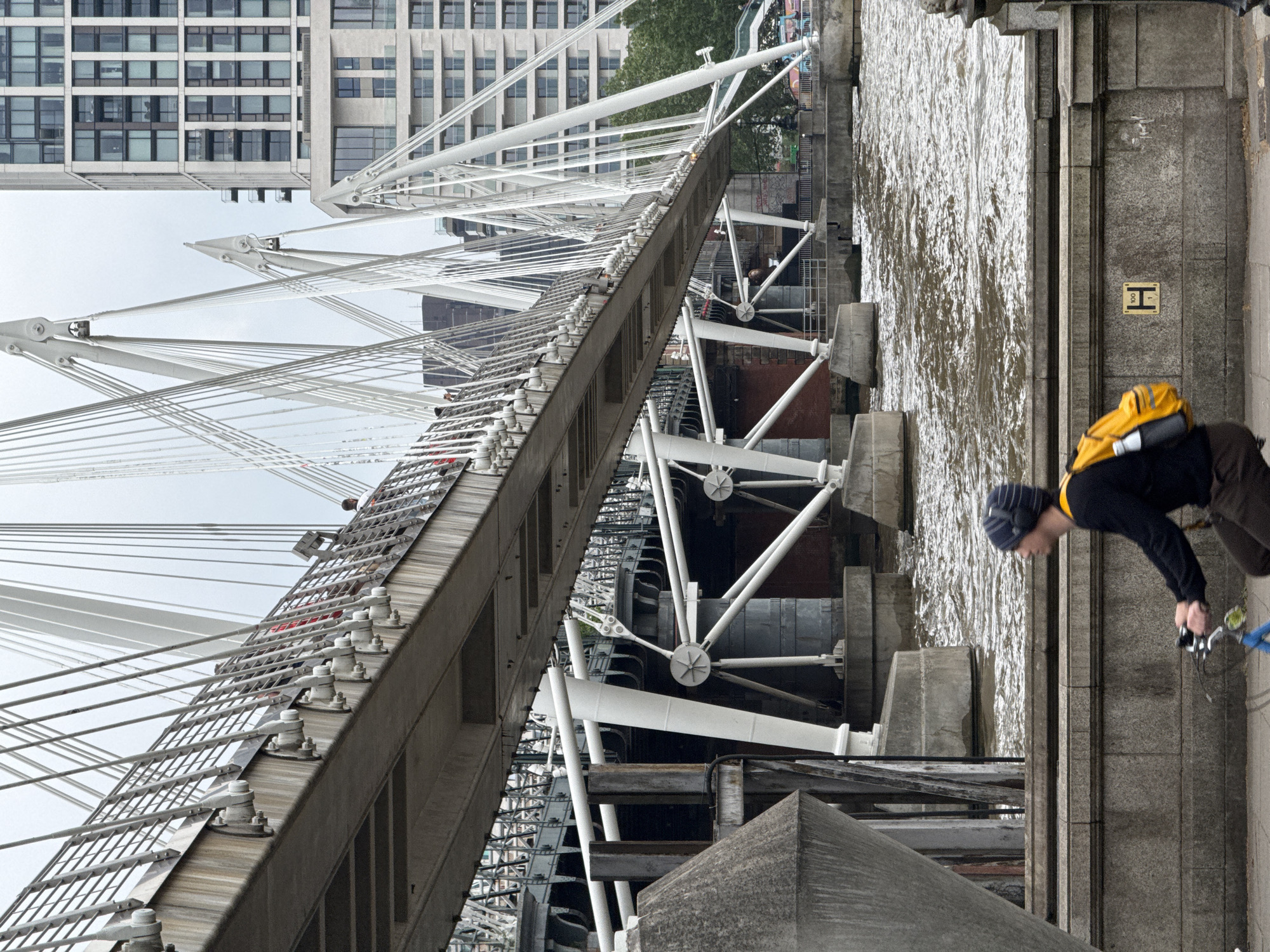 The image depicts the Golden Jubilee Bridge in London, United Kingdom, viewed from a perspective looking along the edge of the bridge towards the water. The structure of the bridge, with its white support cables and steel framework, dominates the left side of the image. A tall building with numerous windows is visible in the background. On the right side of the image is a concrete walkway along the bridge. A man wearing a dark shirt, dark pants, a striped hat, and a yellow backpack is leaning over the edge of the bridge, holding a blue object in his hand. The water of the River Thames is visible below the walkway. A yellow sign with a black "H" is on the side of the walkway.